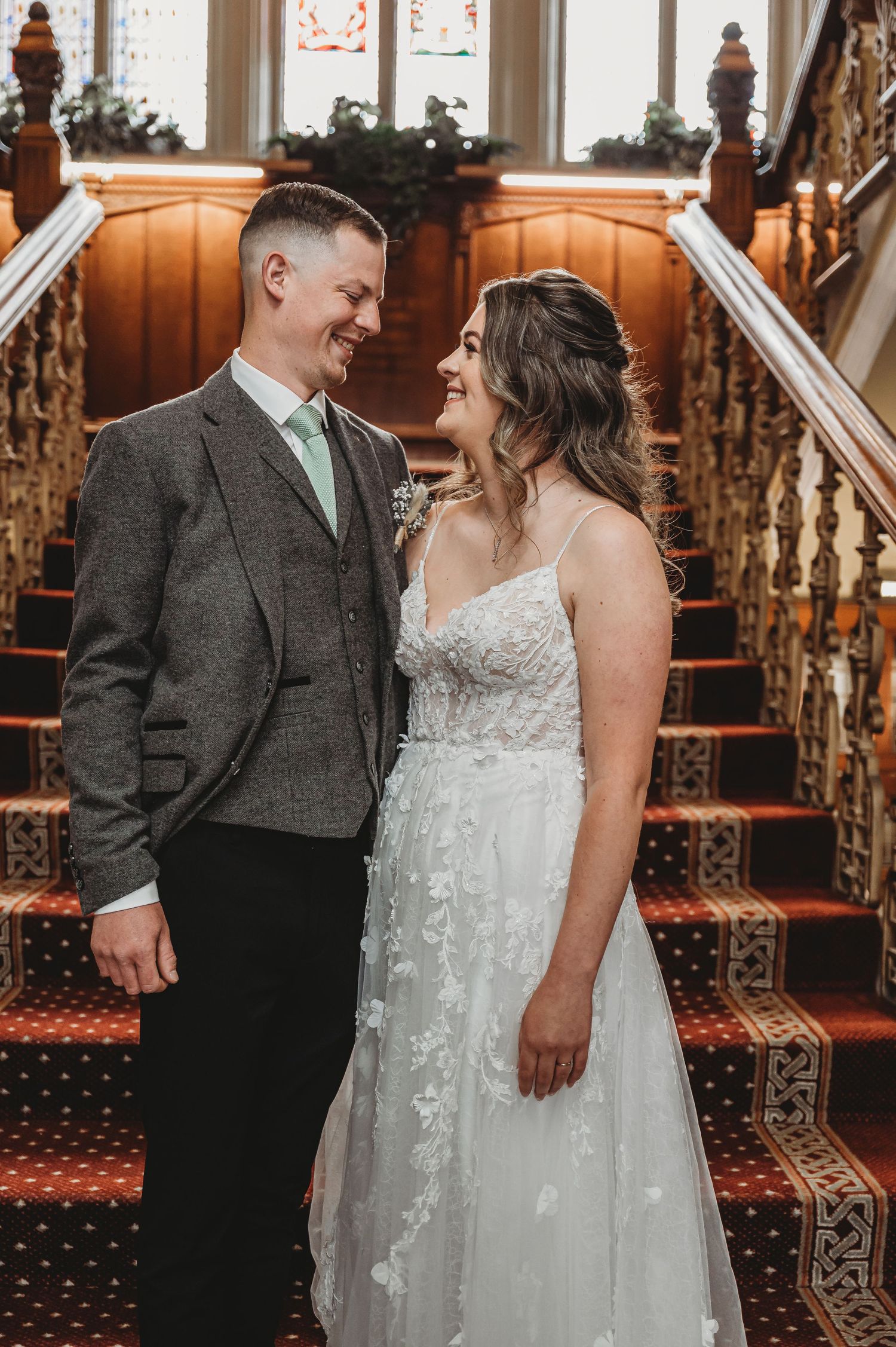 A couple stands on an ornate staircase with red carpeting, wearing wedding attire and sharing an intimate moment.