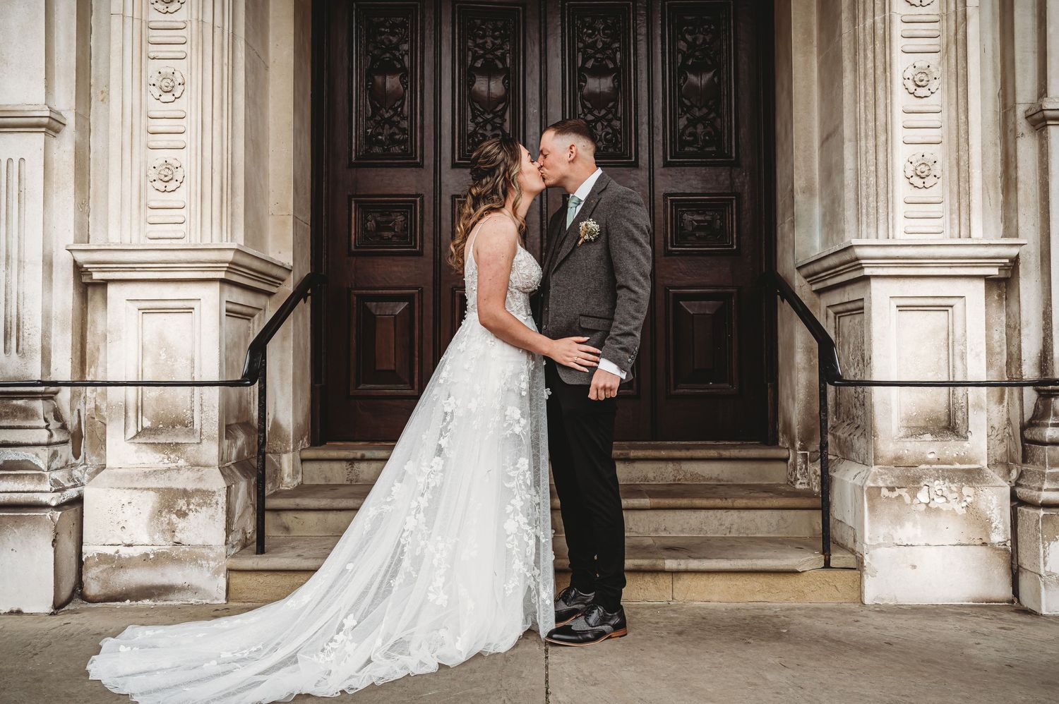 Elegant wedding couple embraces in front of ornate wooden doors at historic stone building entrance.