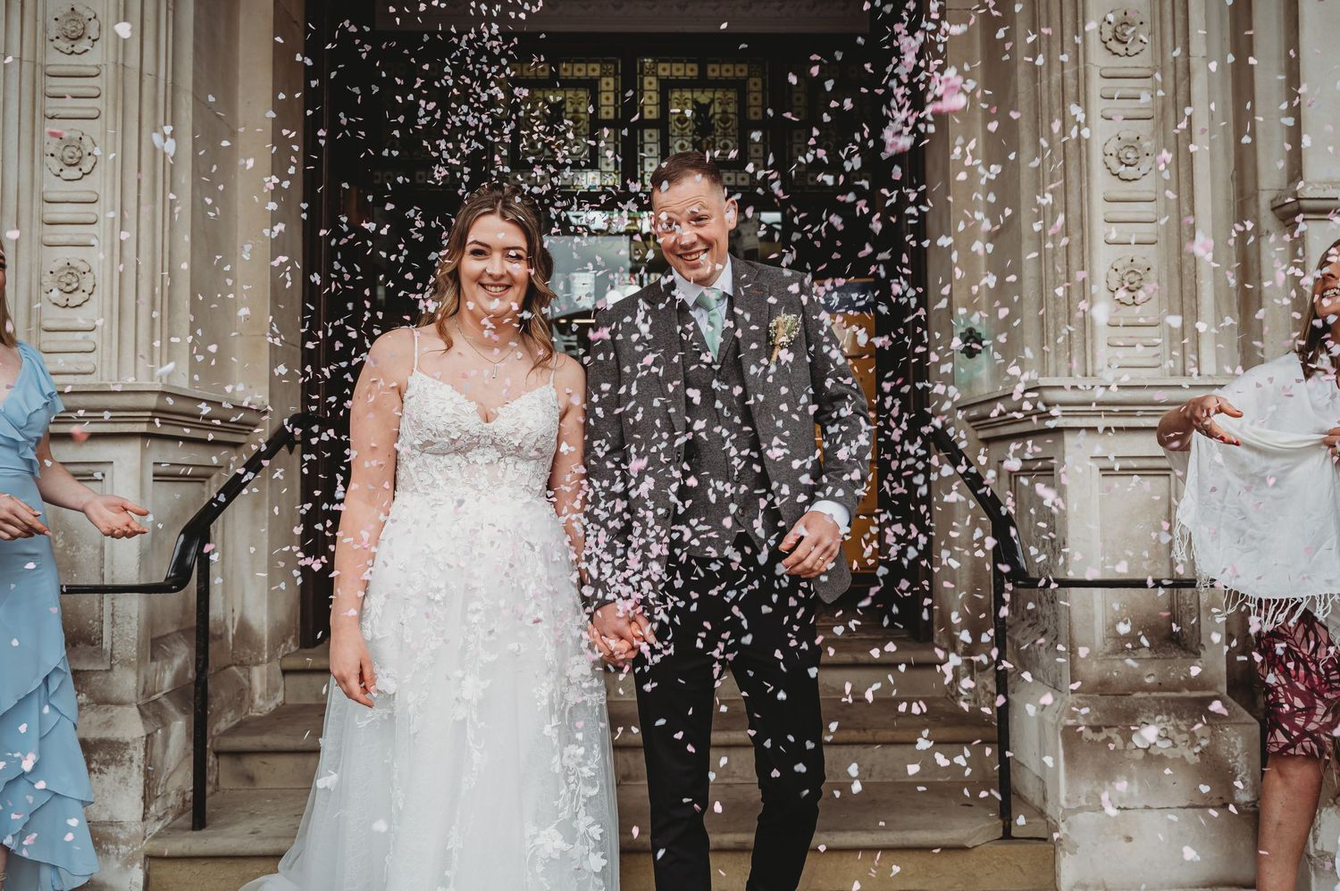 Newlyweds stand at church entrance while guests throw confetti around them during joyful celebration.
