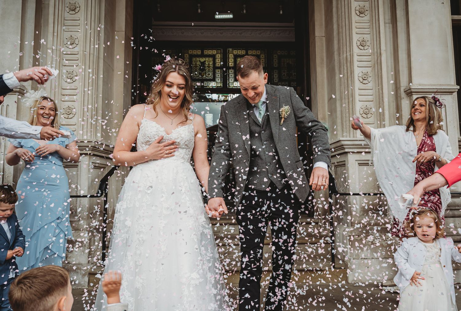 Wedding couple exits church surrounded by falling confetti on stone steps with guests celebrating the joyous moment.