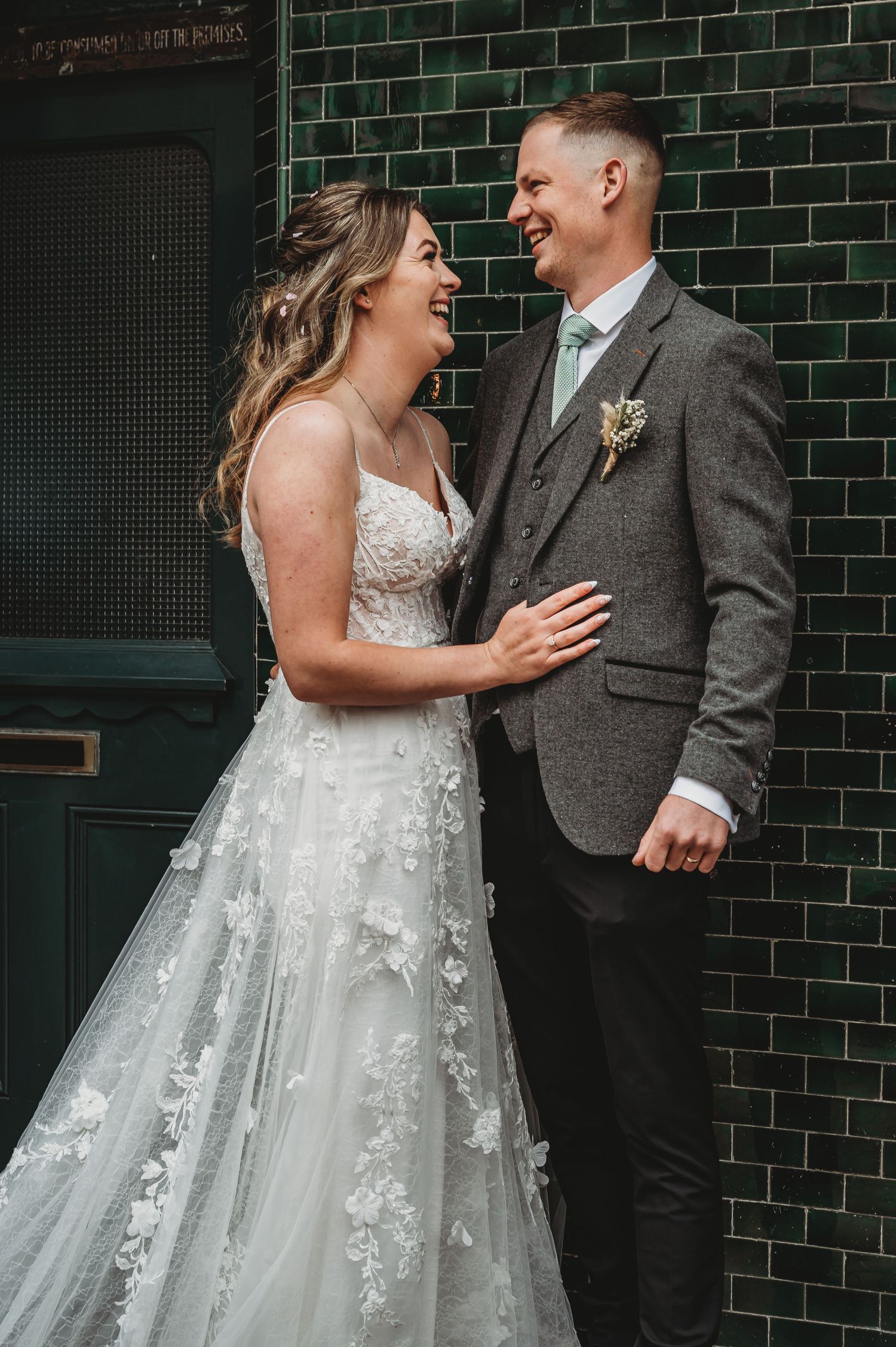 A happy couple shares a candid moment laughing together against a dark green brick wall at their wedding.