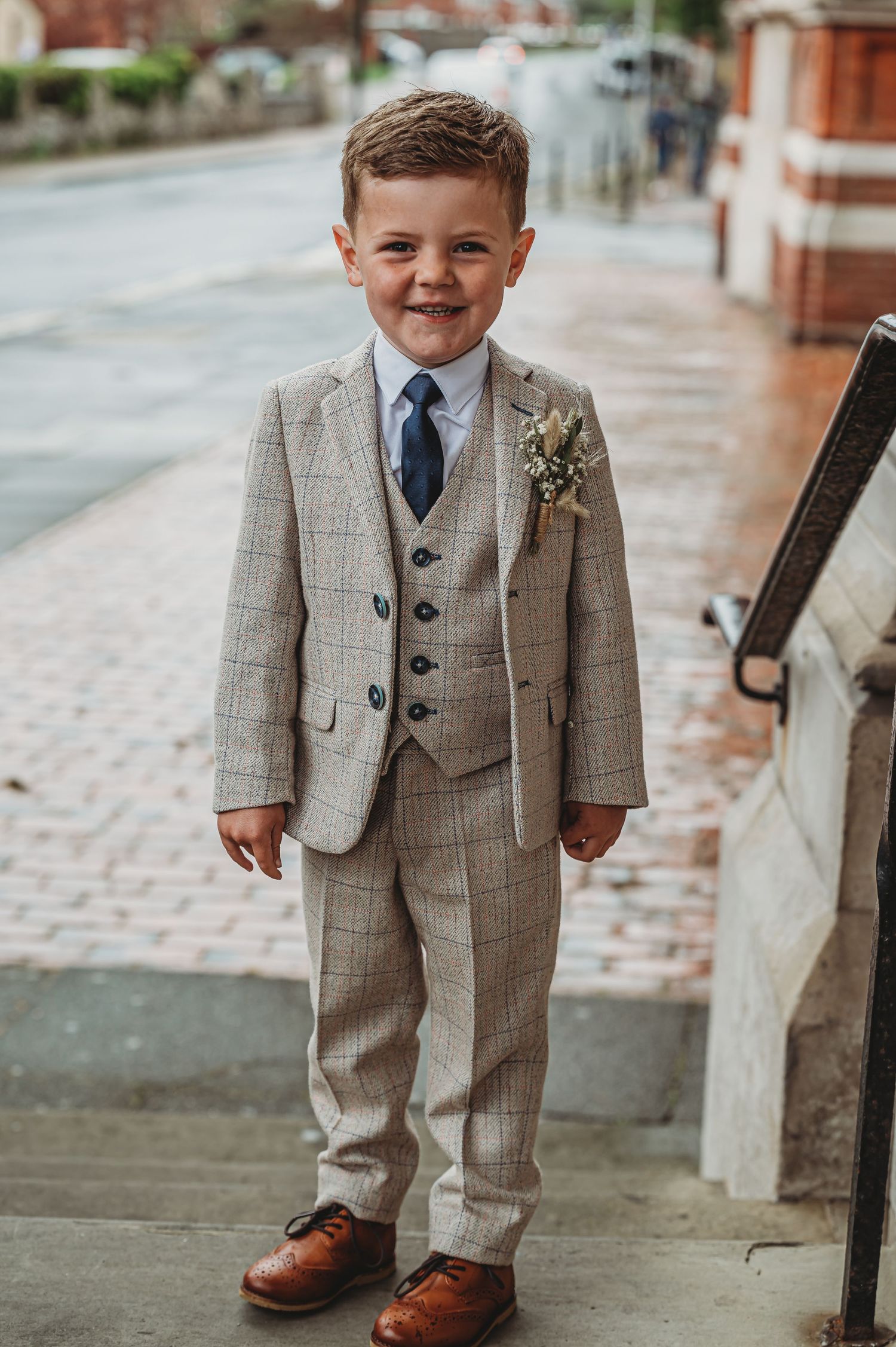 A young person in a well-fitted gray three-piece suit with a blue tie stands on a brick sidewalk for a formal occasion.