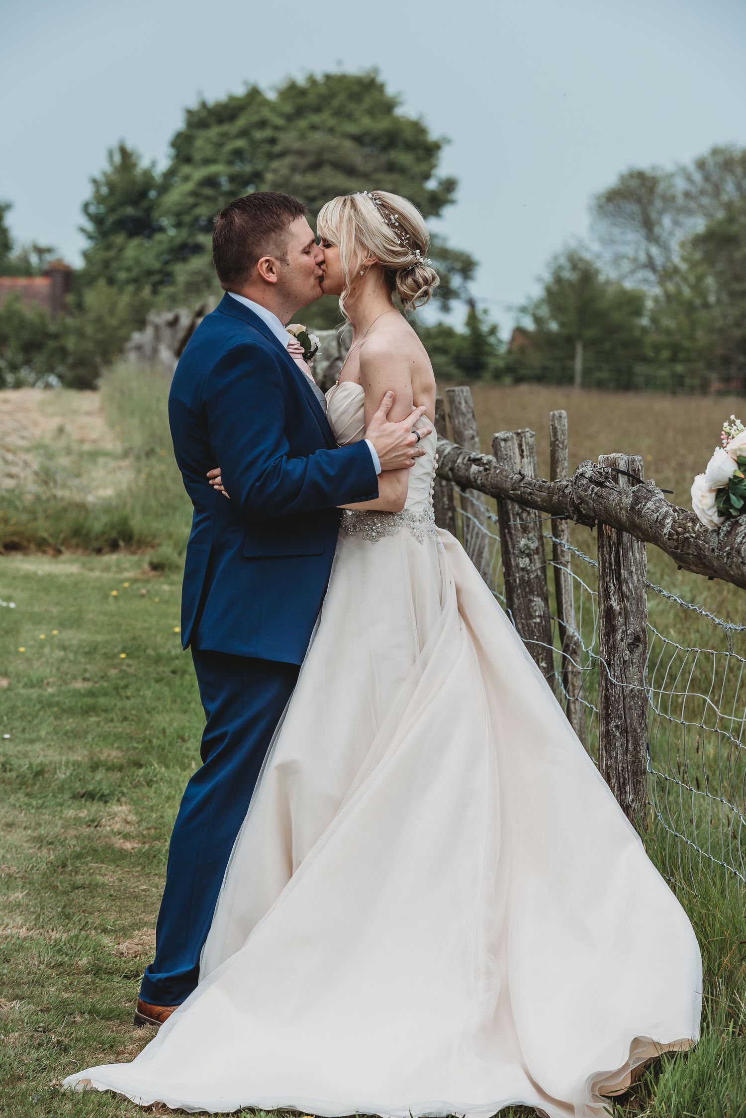A romantic wedding couple shares a kiss beside a rustic wooden fence in an outdoor countryside setting.
