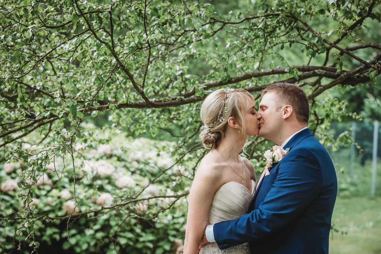 A romantic wedding couple shares a tender kiss under flowering spring tree branches in a garden setting.