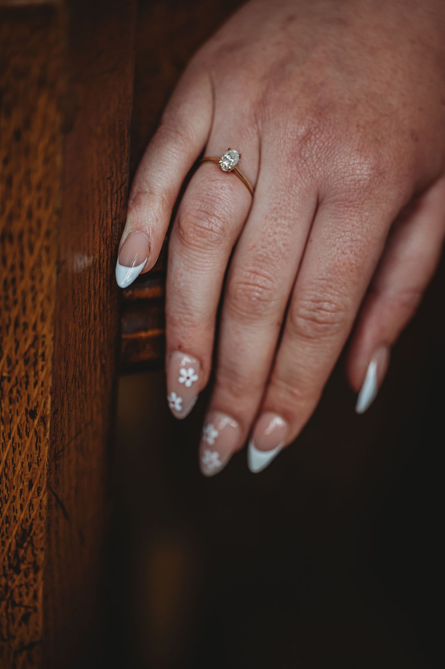 Close-up of French manicured nails with delicate floral designs and a dainty solitaire ring on wooden background.