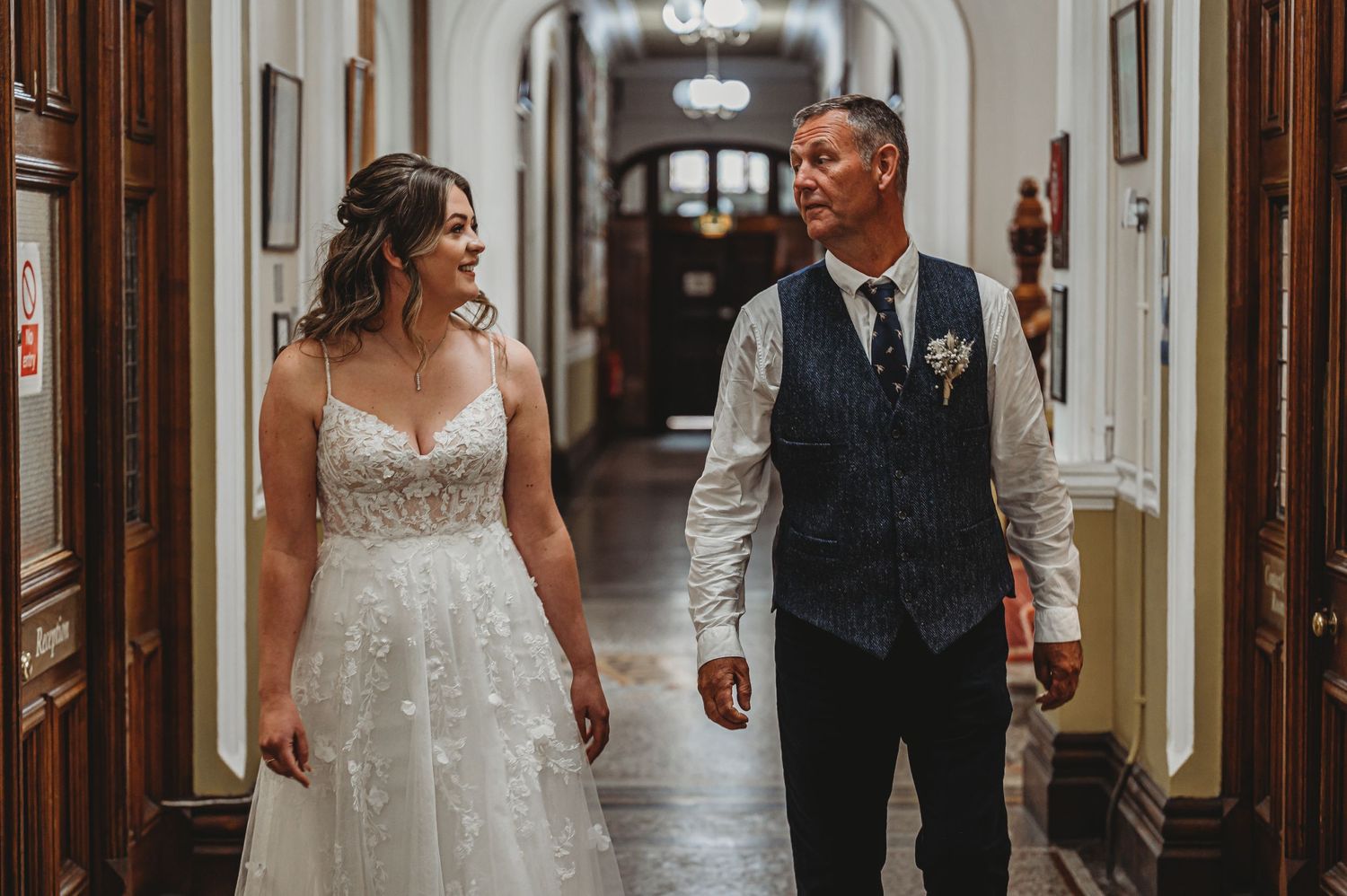 A couple in wedding attire walking together down an elegant hallway with ornate walls and lighting fixtures.