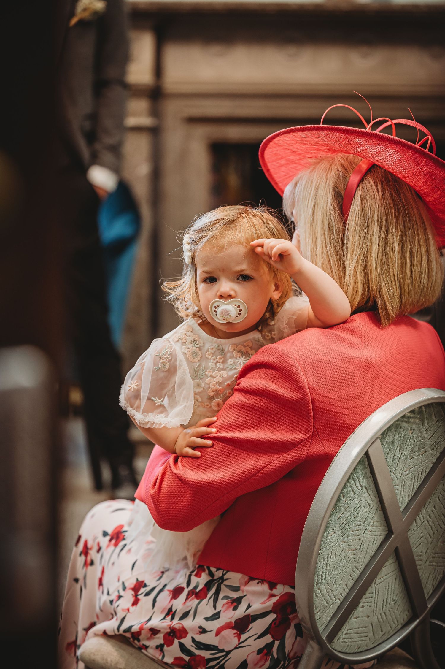 A small child with a pacifier being held by someone wearing a bright red hat and floral dress.