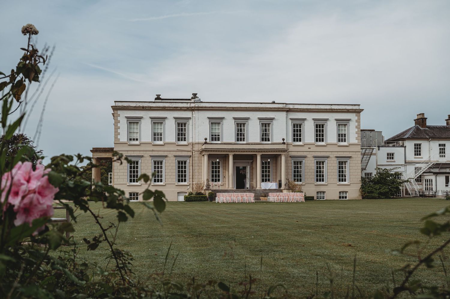 Elegant Georgian mansion with white facade and columns stands on manicured lawn with pink roses in foreground.