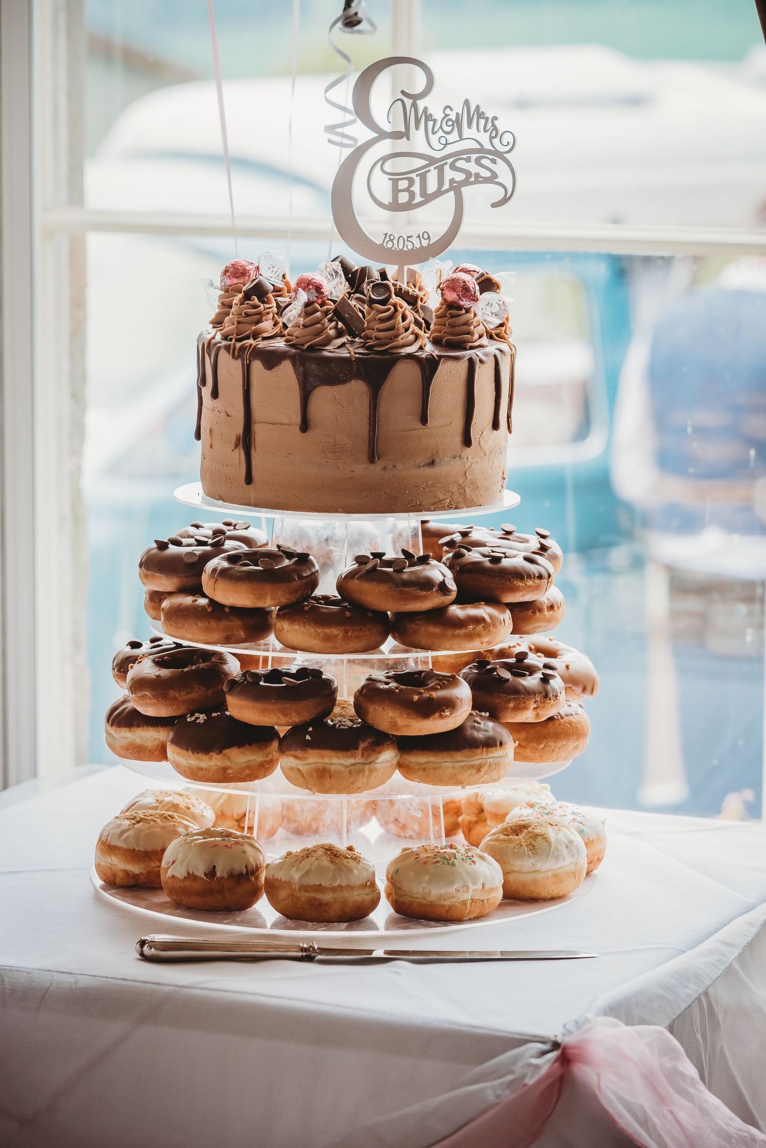 A wedding dessert tower featuring a chocolate drip cake on top and tiers of decorated donuts below on a white stand.