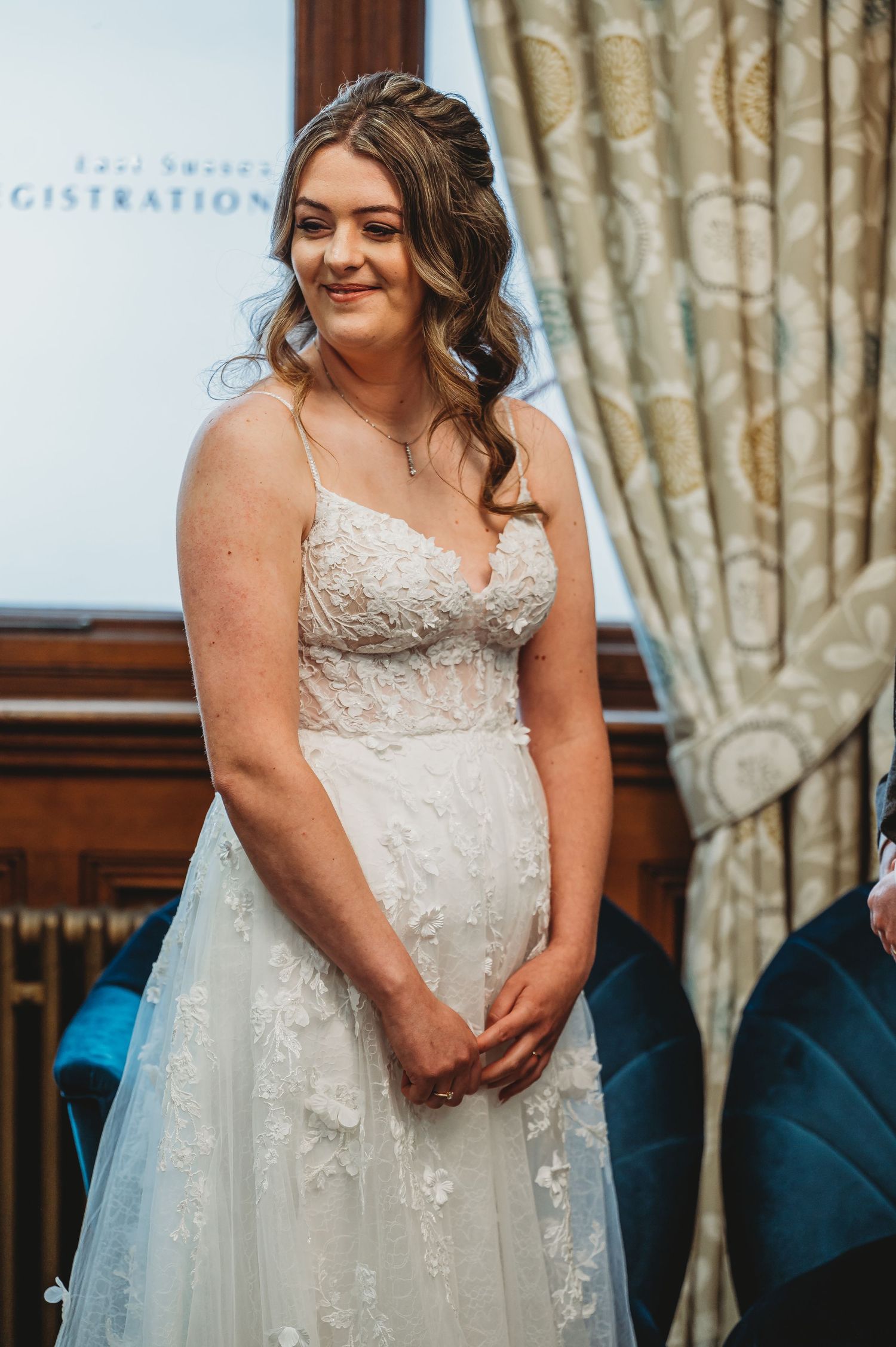 A bride wearing a white lace wedding dress with spaghetti straps smiles during her wedding ceremony.