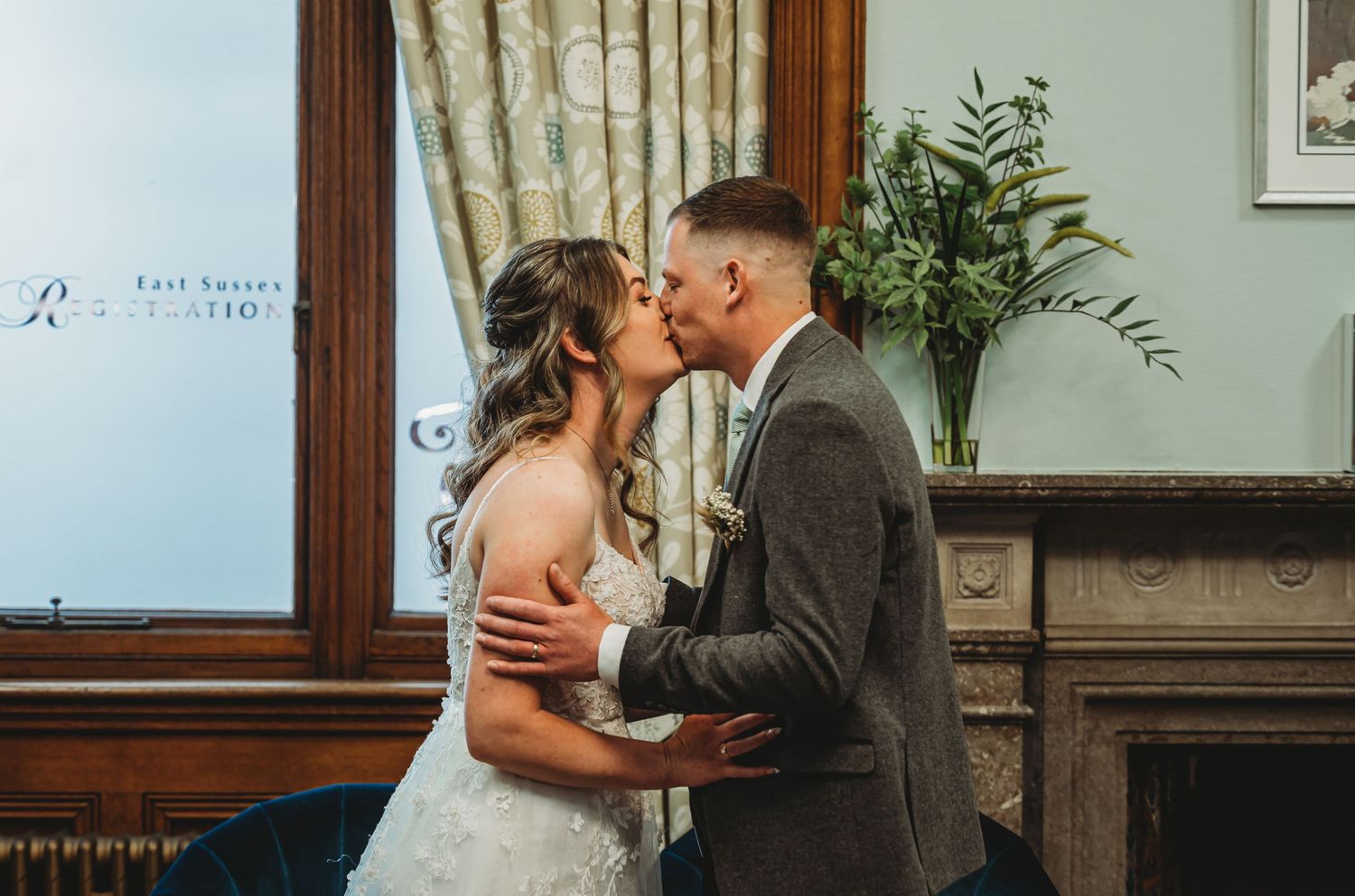 A romantic wedding couple share their first kiss in an elegant indoor venue next to a vintage fireplace.