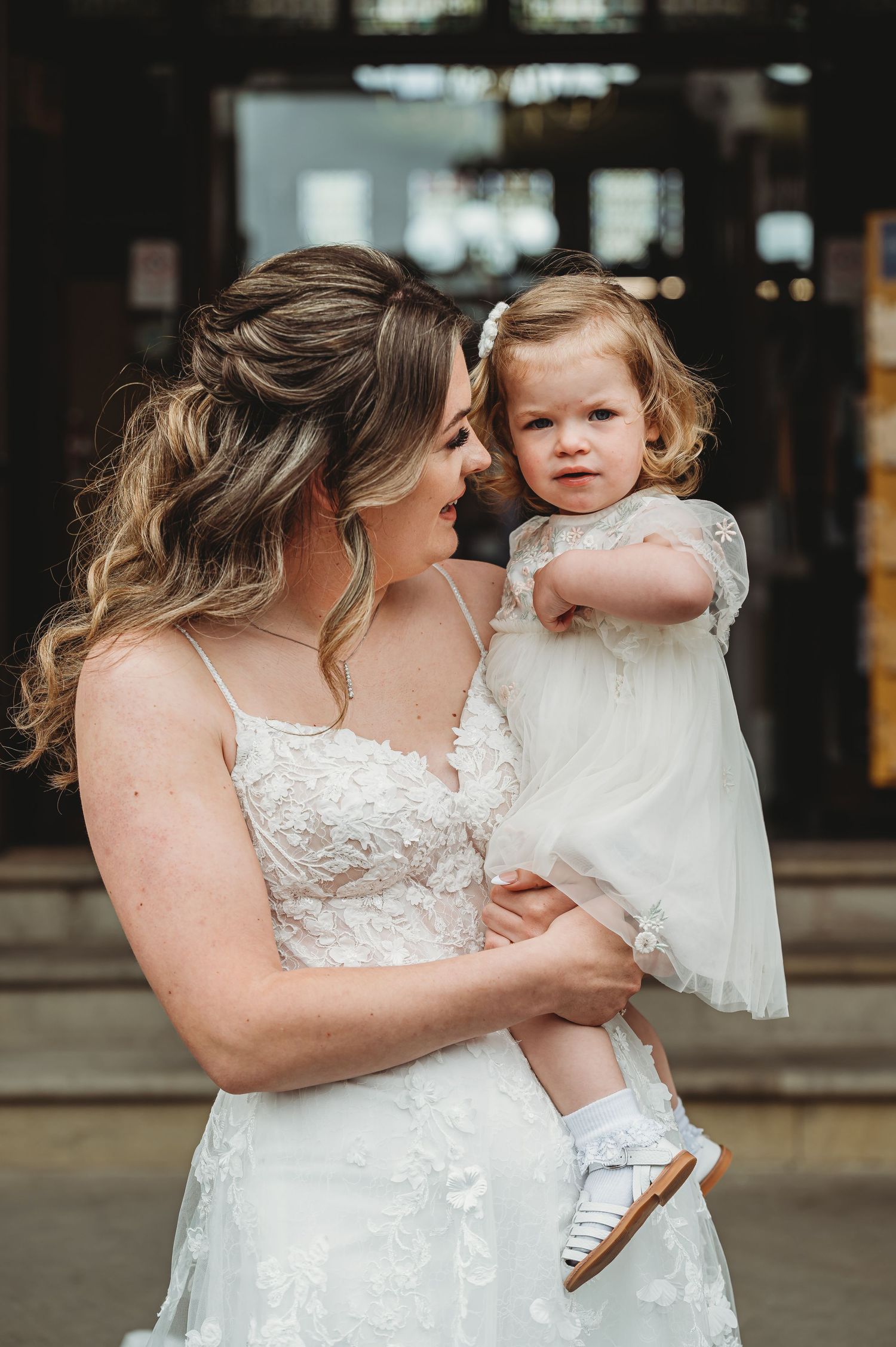 A bride in a lace wedding dress holds a small child wearing a white dress at an indoor venue.