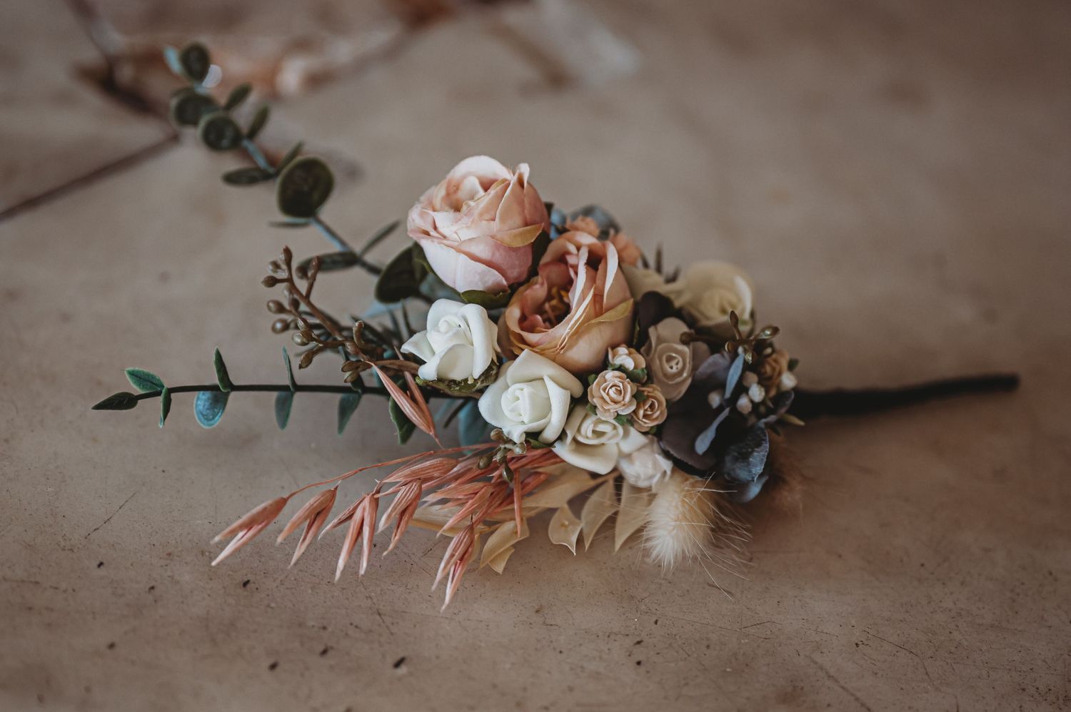 A romantic wedding boutonniere with peach roses, dried flowers, and eucalyptus on a matte textured surface.