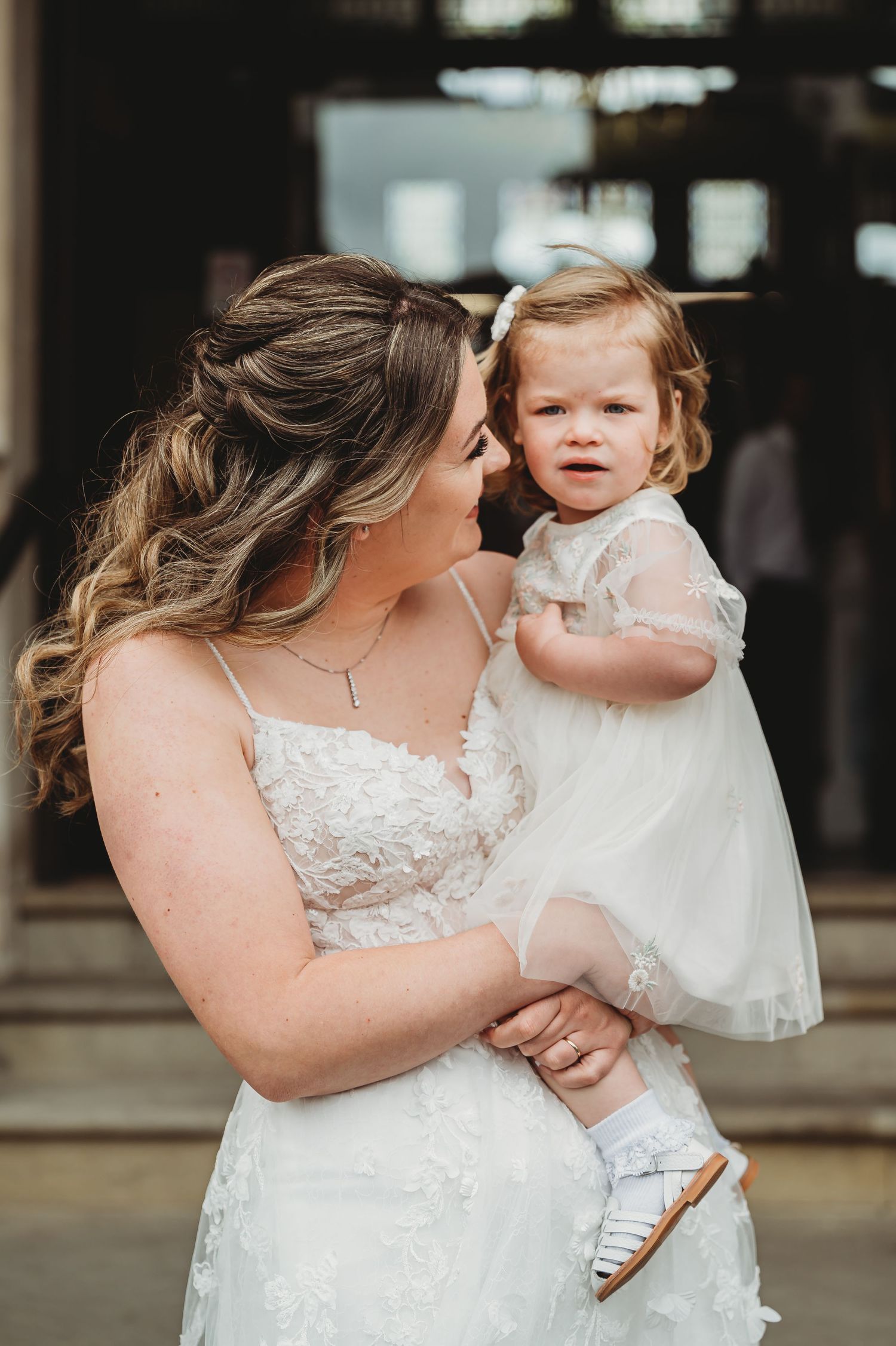 A tender candid moment between woman in white lace wedding dress holding a small child wearing a white tulle dress.