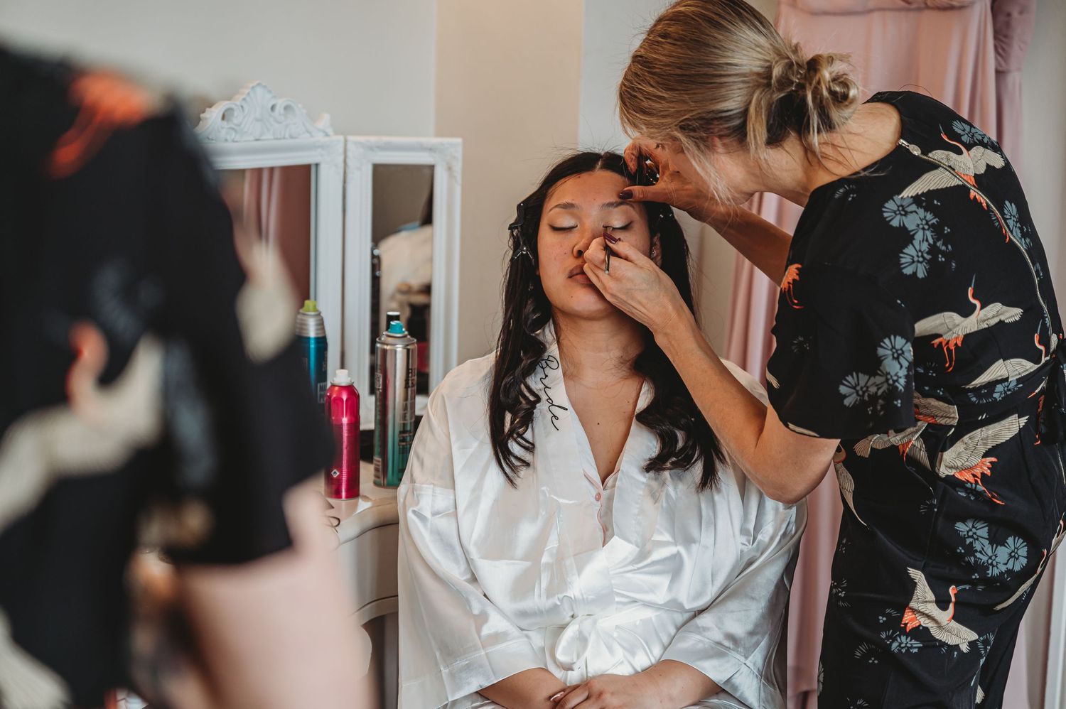 A makeup artist applies cosmetics to someone wearing a white silk robe during a beauty preparation session.