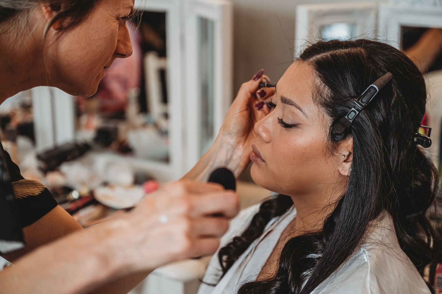 A makeup artist carefully applies cosmetics to a model with long dark hair wearing hair clips while sitting in a brightly lit room.