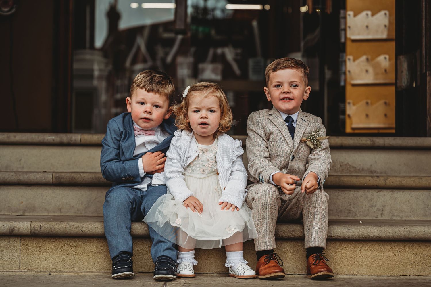 Three young children in formal attire sit together on outdoor stone steps.