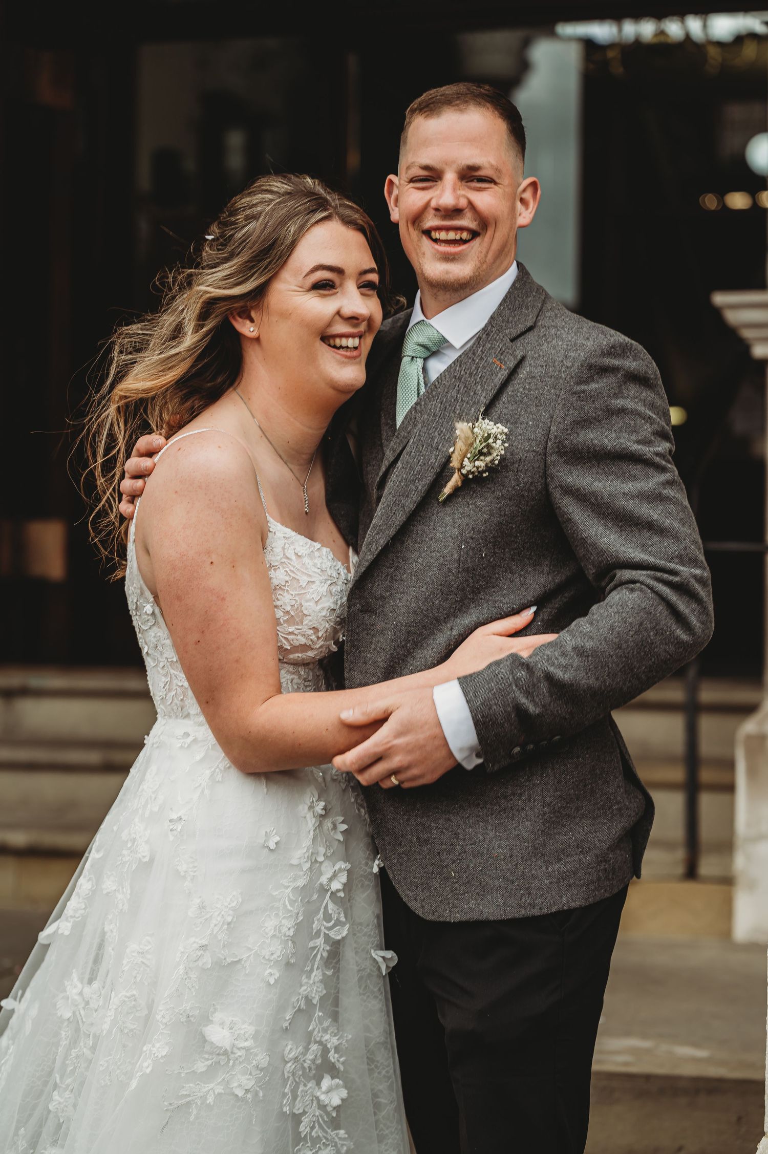 A joyful couple shares a candid moment on their wedding day, the bride in white lace and groom in gray tweed.