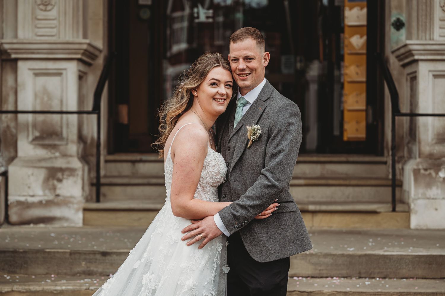 A newlywed couple embraces and laughs together on their wedding day outside a grand stone building entrance.