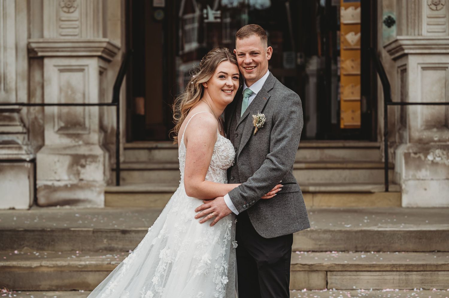 A happy couple shares a romantic moment on stone steps outside an elegant historic building in wedding attire.