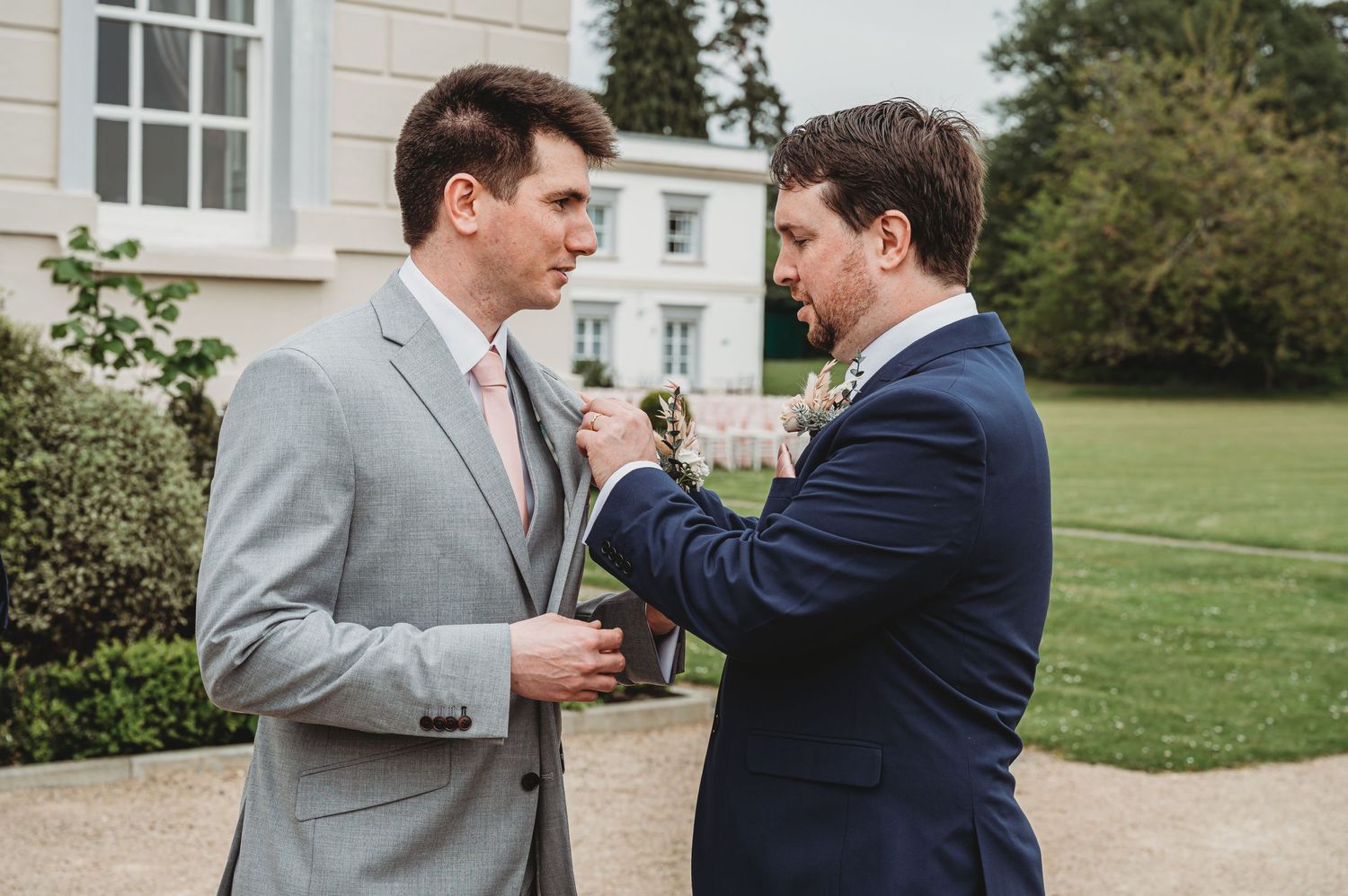 Two men in suits share a moment at an outdoor wedding, with one adjusting the other's boutonniere.