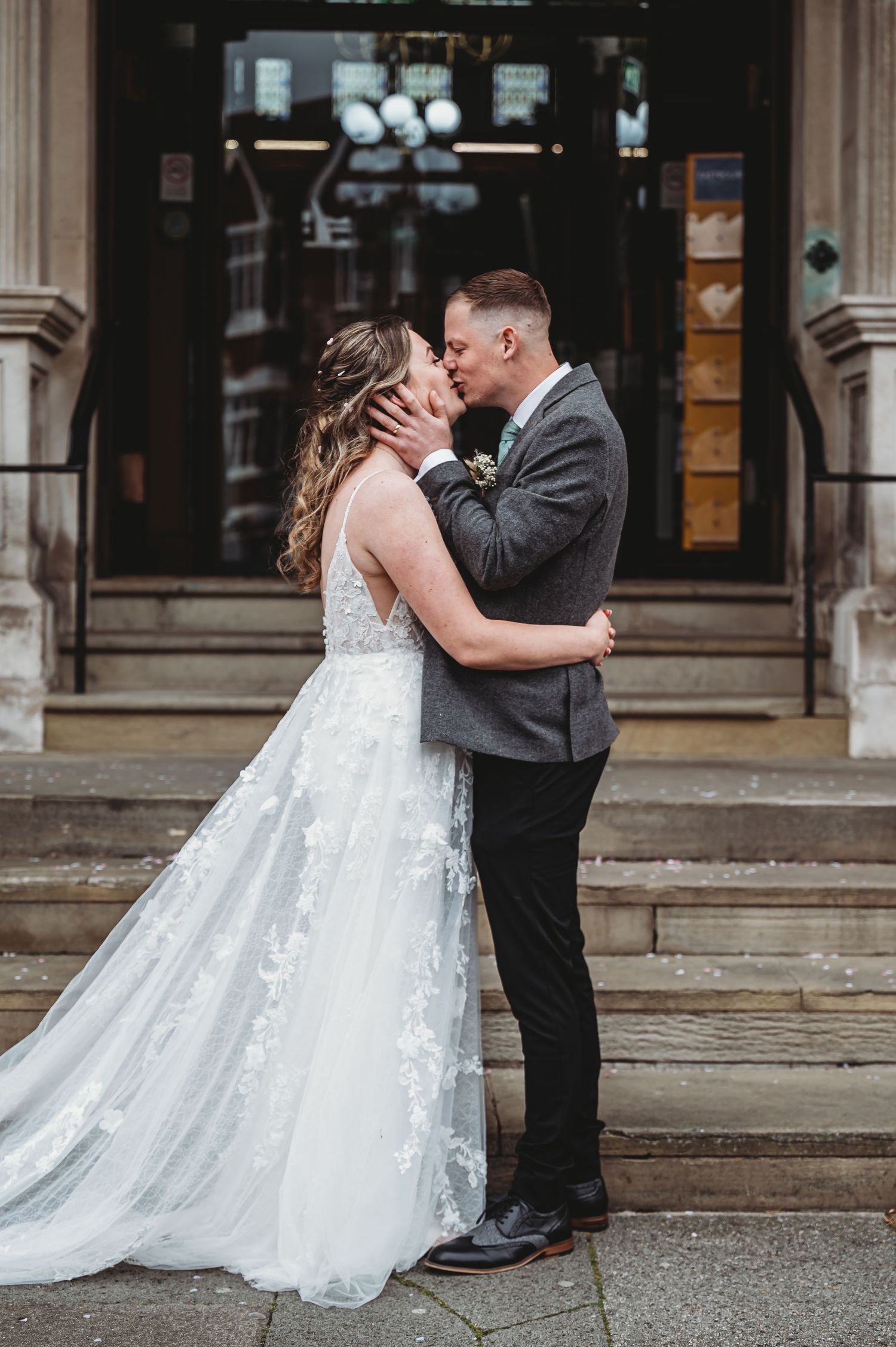 A romantic wedding couple sharing a kiss on stone steps in front of a historic building entrance.