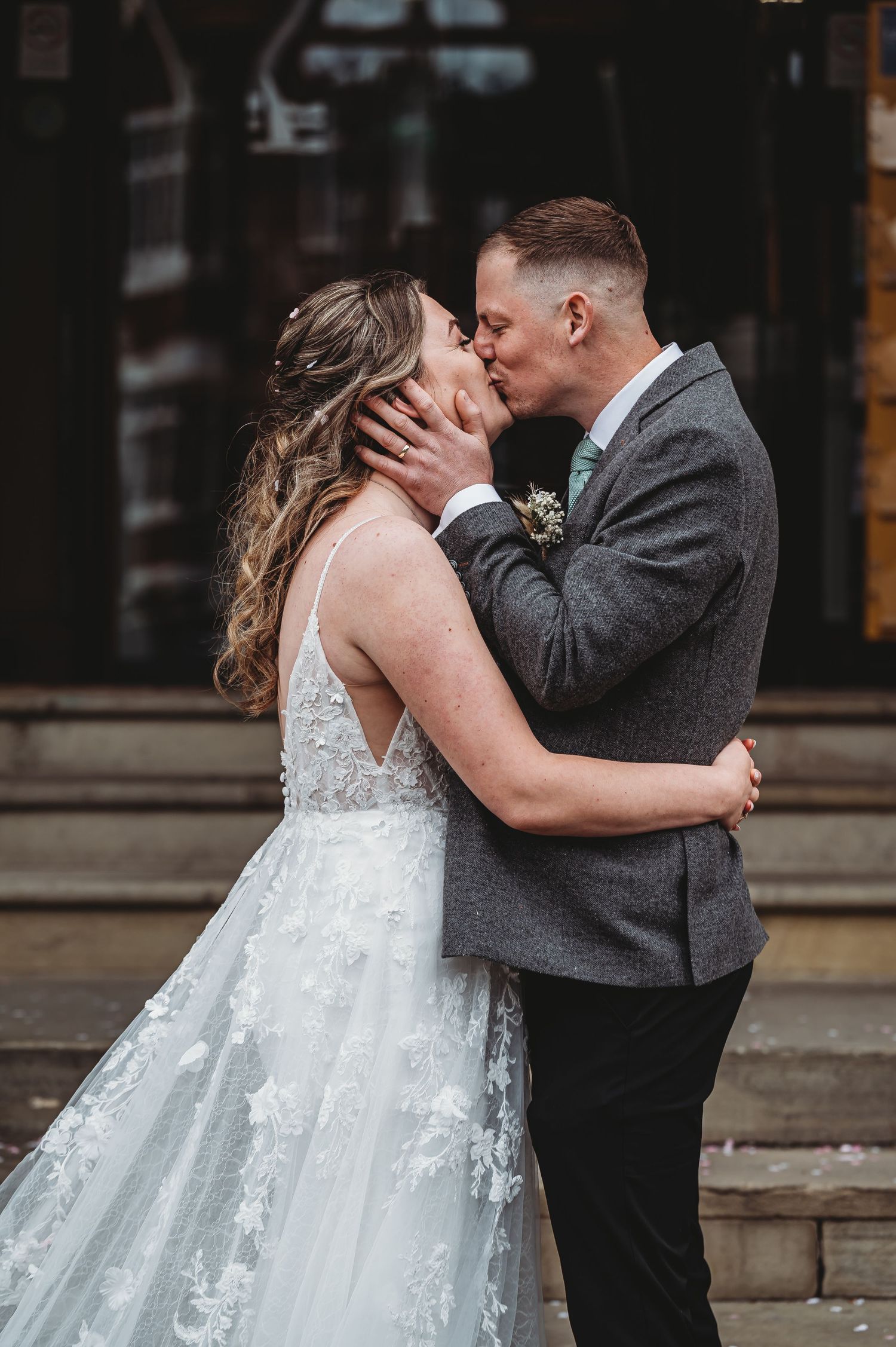 A couple shares a romantic kiss on their wedding day, the bride wearing a lace gown and groom in a grey suit.