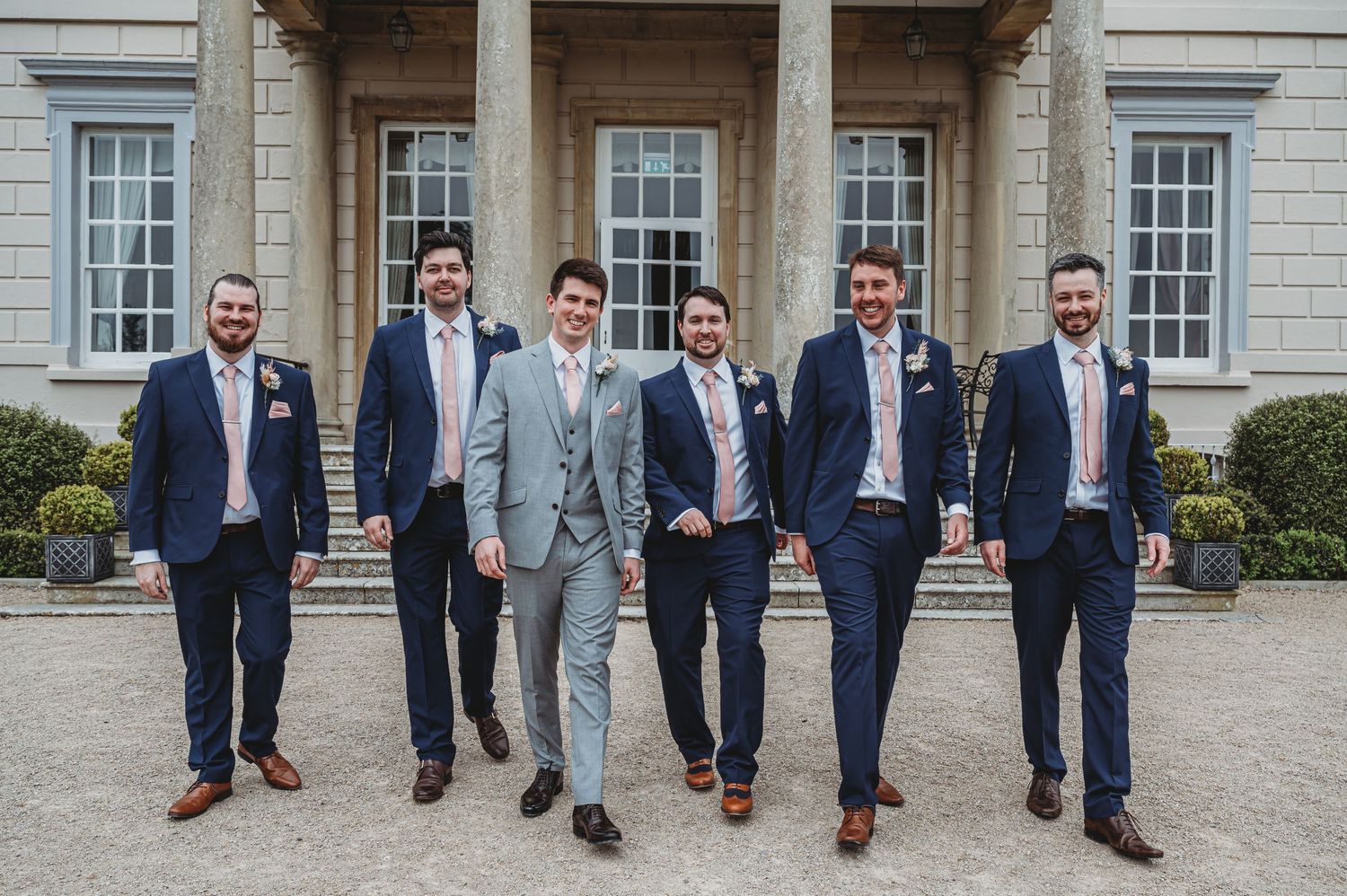 Groomsmen in coordinated navy and light gray suits stand together outside an elegant stone building with columns.