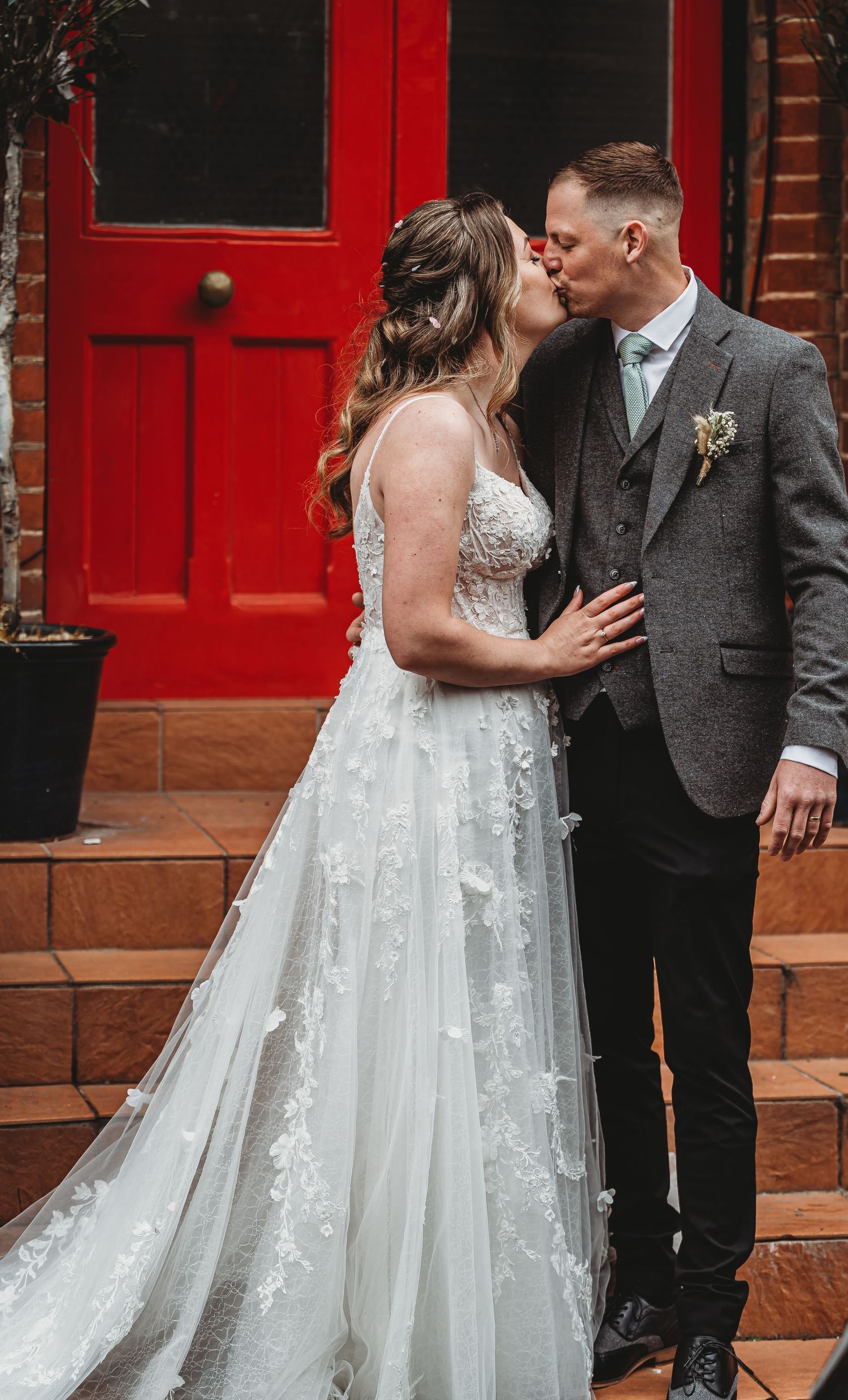 A newlywed couple kissing in front of a vibrant red door, with the bride wearing a detailed lace wedding gown.