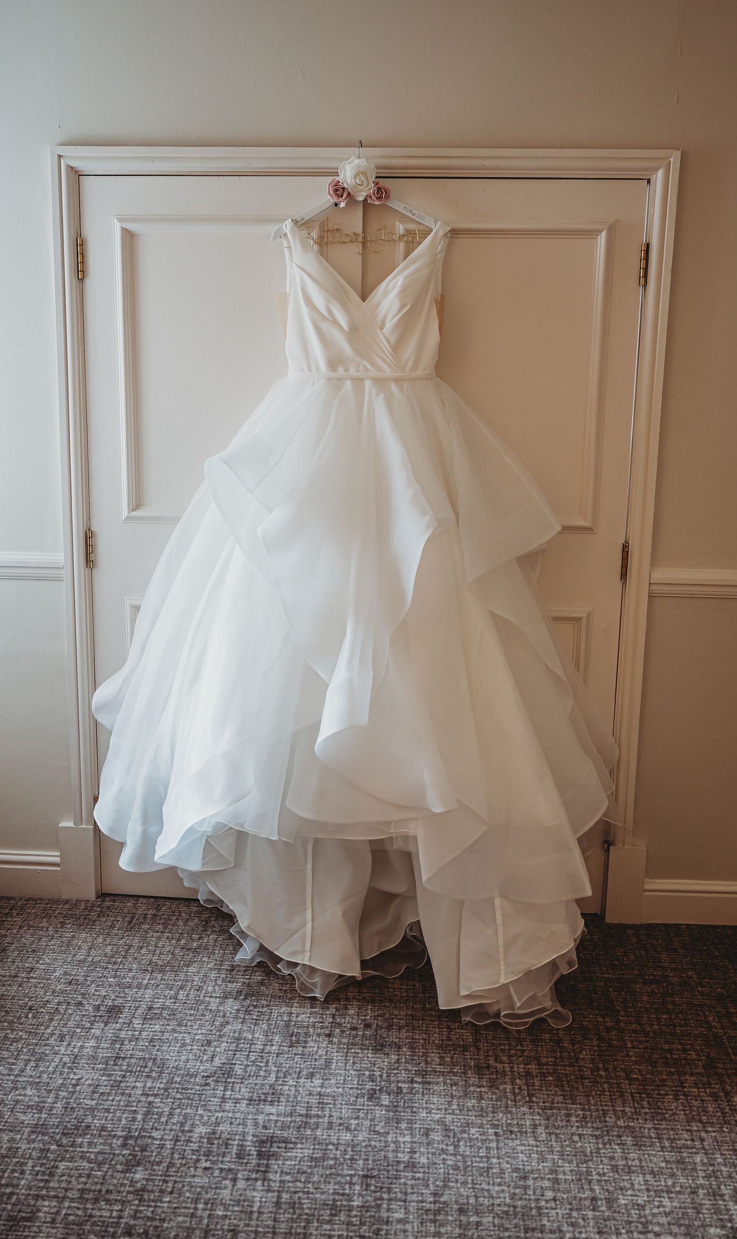 An elegant white wedding dress with a v-neck bodice and flowing ruffled skirt hangs on display against a paneled wall.