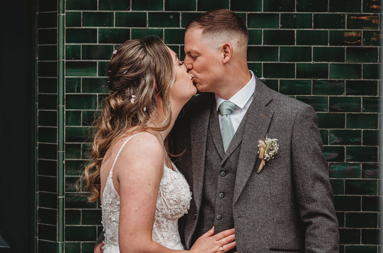Newlywed couple sharing intimate kiss against emerald green tiled wall, with groom in grey suit and small boutonniere.