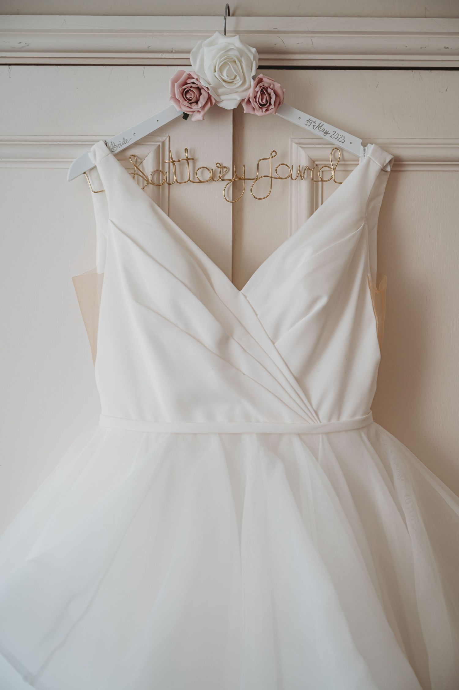 A white wedding dress with a v-neck bodice hangs on a customized hanger adorned with vintage pink and white roses.