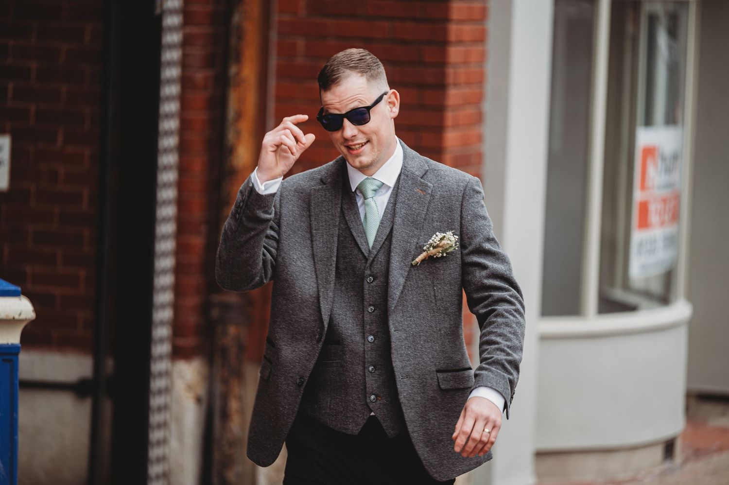 A well-dressed gentleman in a grey three-piece suit and sunglasses adjusts his glasses while walking on a sidewalk.
