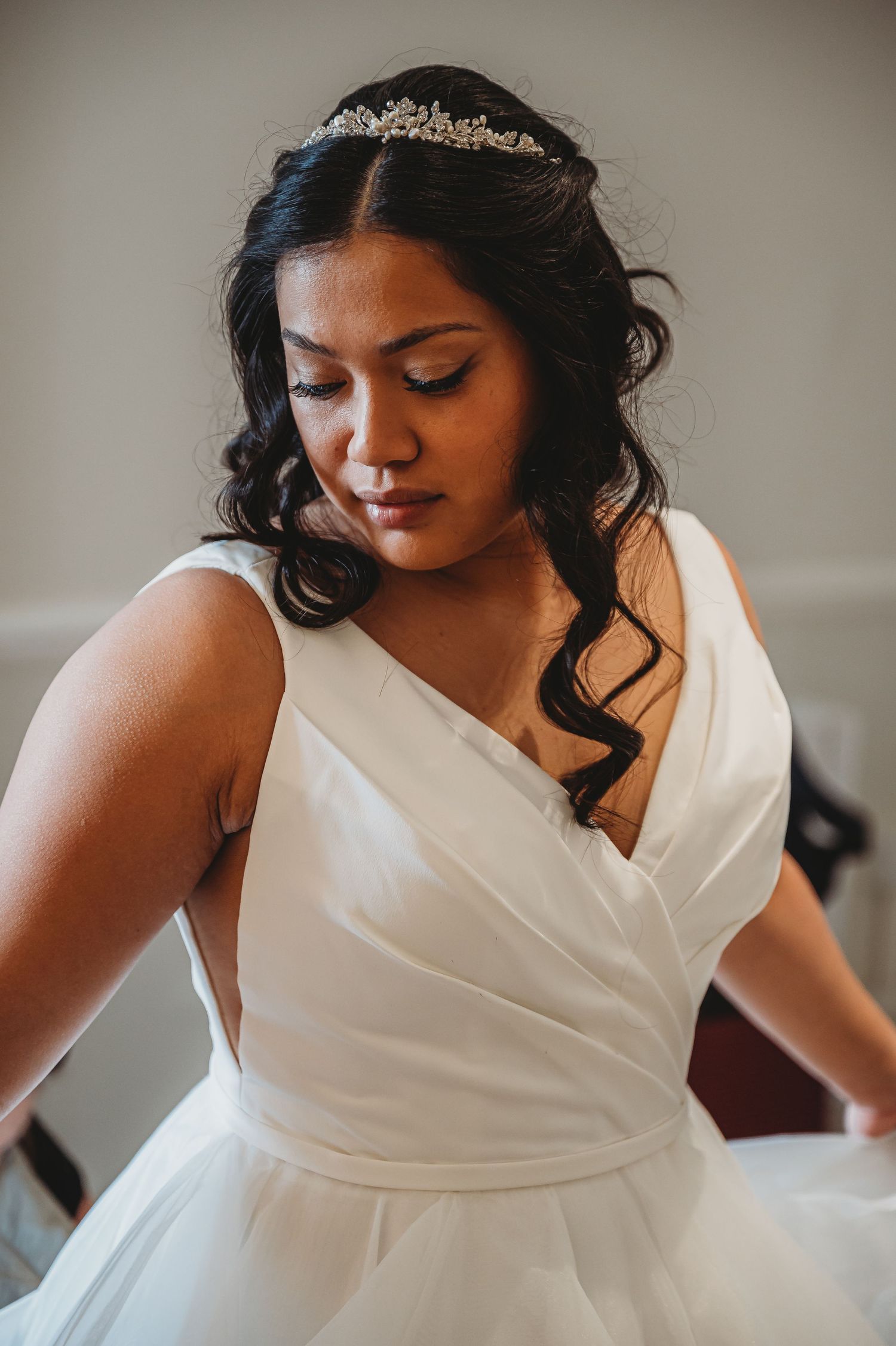 A bride in a classic white wedding dress with a crystal tiara and flowing dark curls gazes down in a lovely portrait.