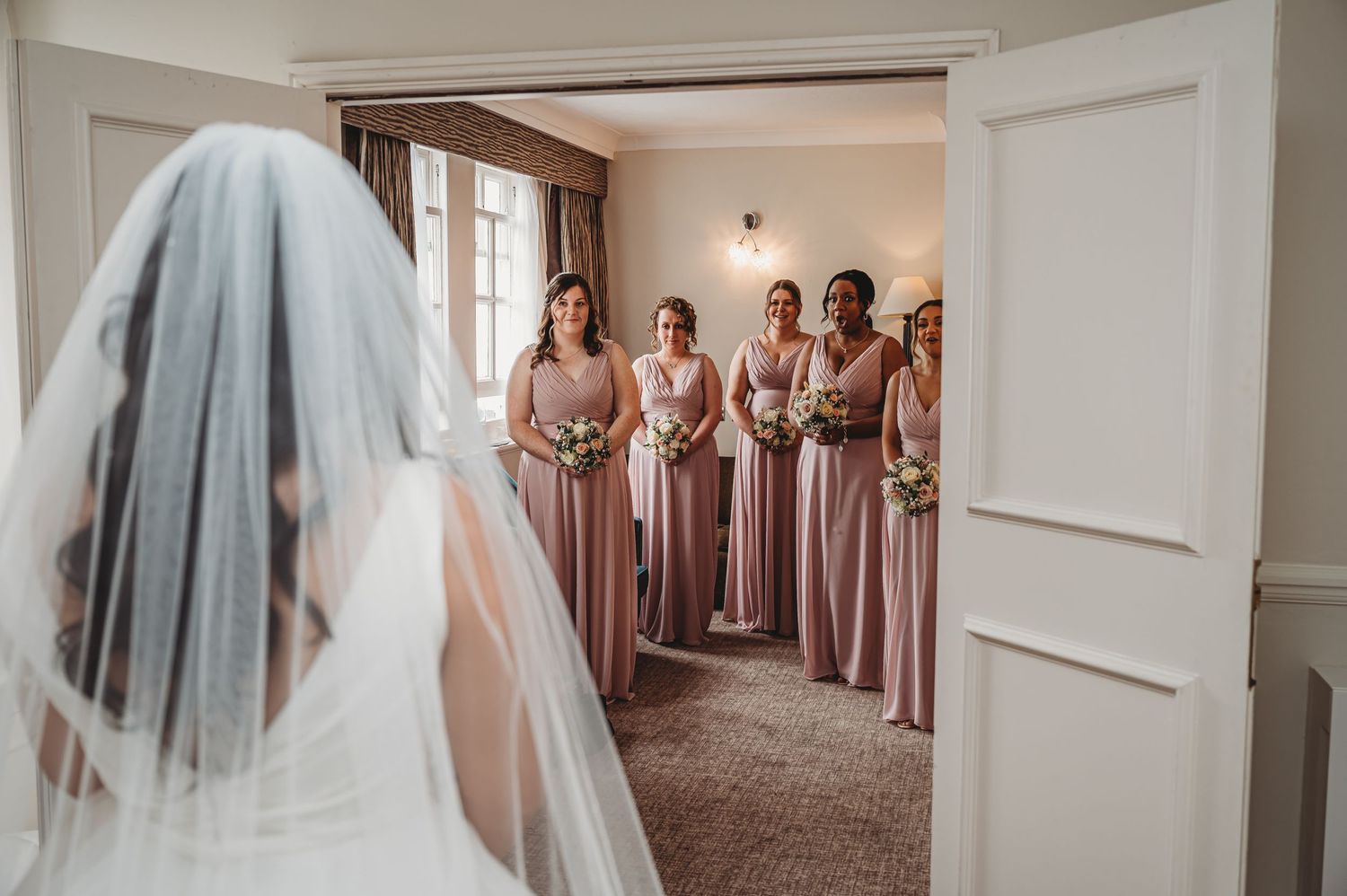 Bridesmaids in blush pink dresses smile as they watch bride in white veil through doorway at wedding venue.