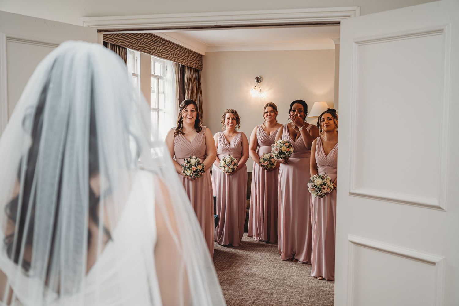 Bridesmaids in dusty rose gowns gather at doorway to greet bride wearing white veil during wedding preparations.