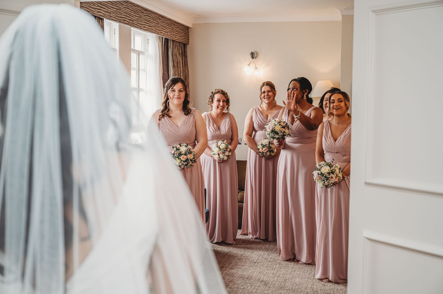 Bridesmaids in dusty rose dresses stand together holding bouquets as they watch the bride in her wedding gown and veil.