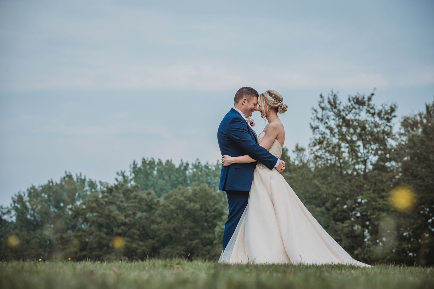 A couple in wedding attire embrace in a scenic meadow with trees in the background against a soft blue sky.