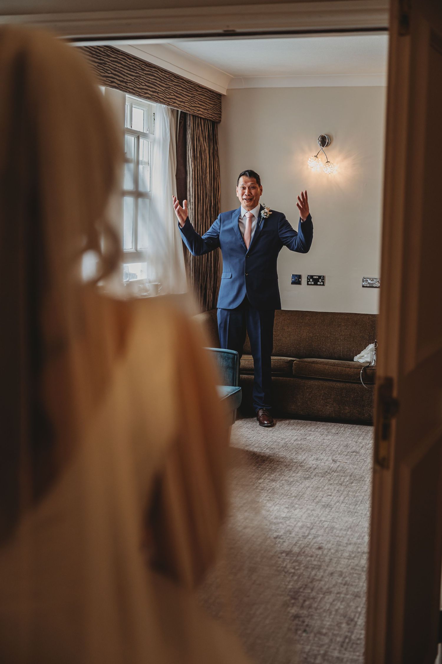 Someone in a navy blue suit reacts with raised hands while standing in an elegant hotel room with draped windows.