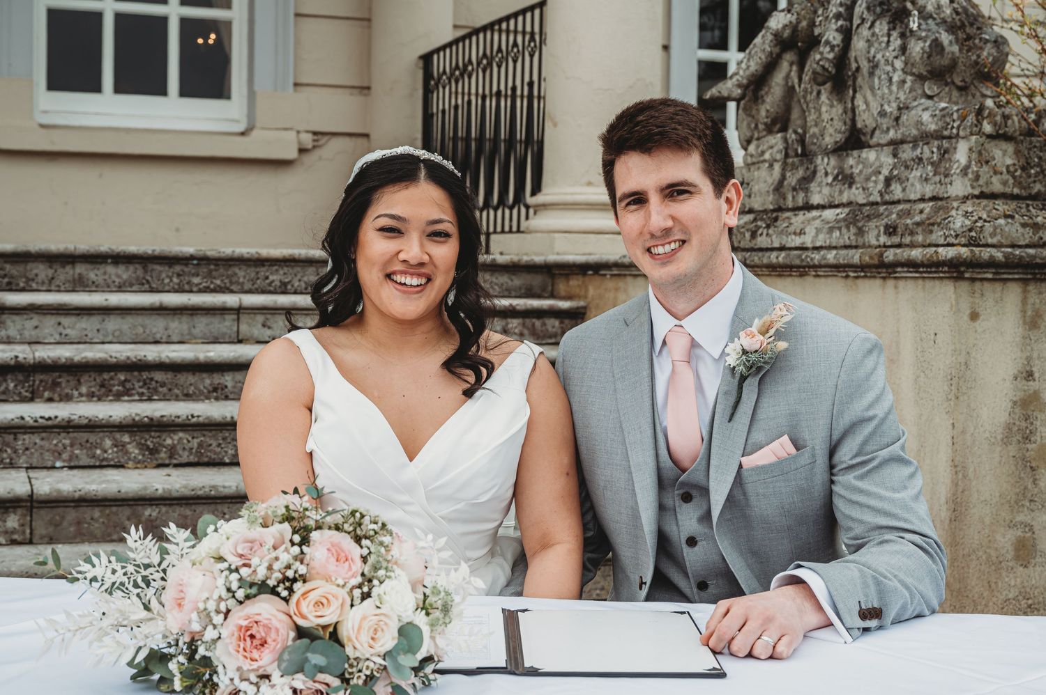 A couple in wedding attire signs marriage documents while seated on elegant stone steps.