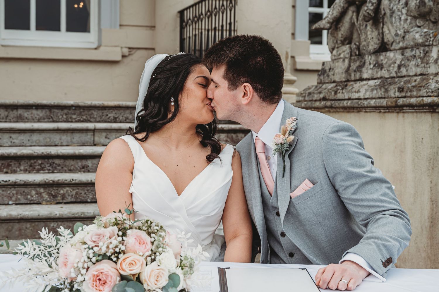 Romantic newlywed couple shares tender moment on stone steps with pink and white floral bouquet.