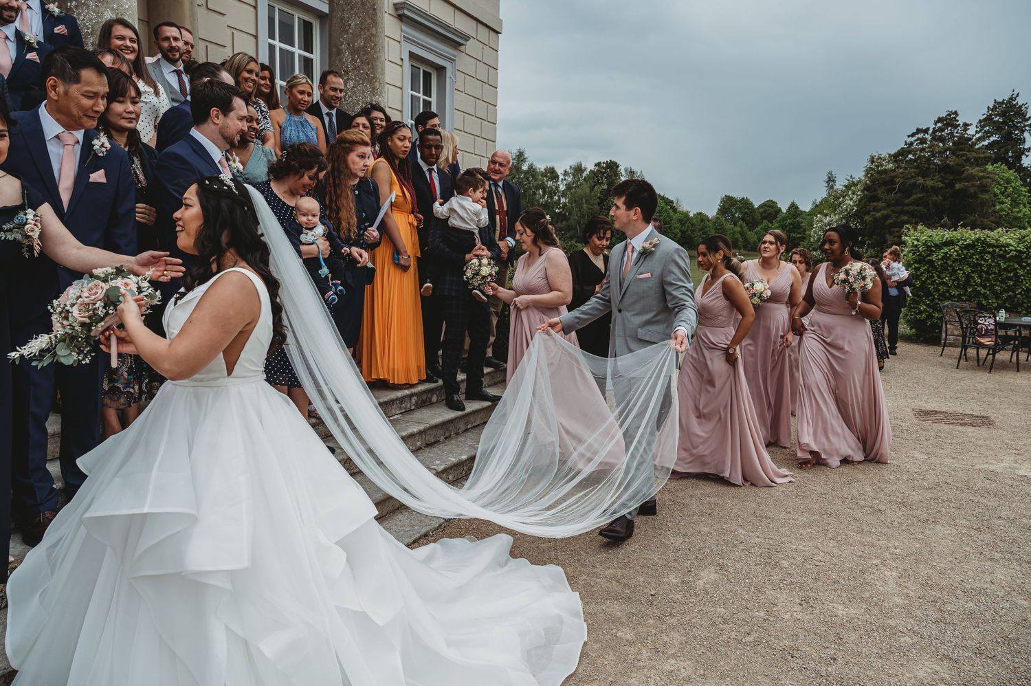 Wedding party gathers outside a stone building as a bride in white gown with flowing veil poses with bridesmaids in pink dresses.