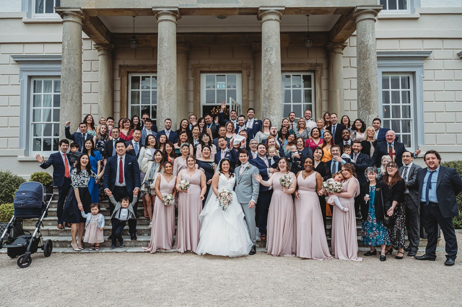 Large wedding party gathered on grand stone steps of an elegant classical building with white pillars and windows.