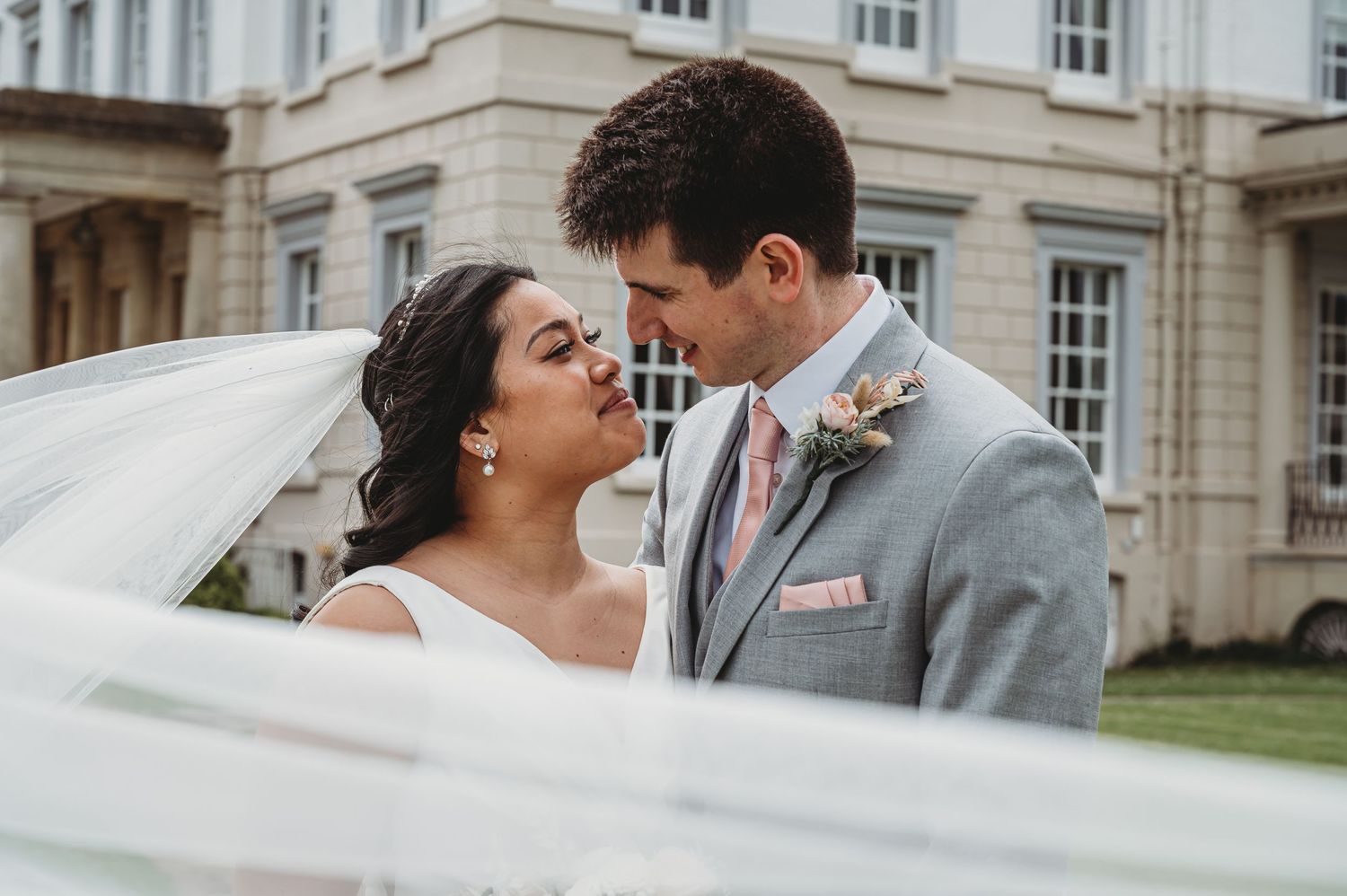 A bride and groom share an intimate moment in front of an elegant historic building as her veil flows in the breeze.