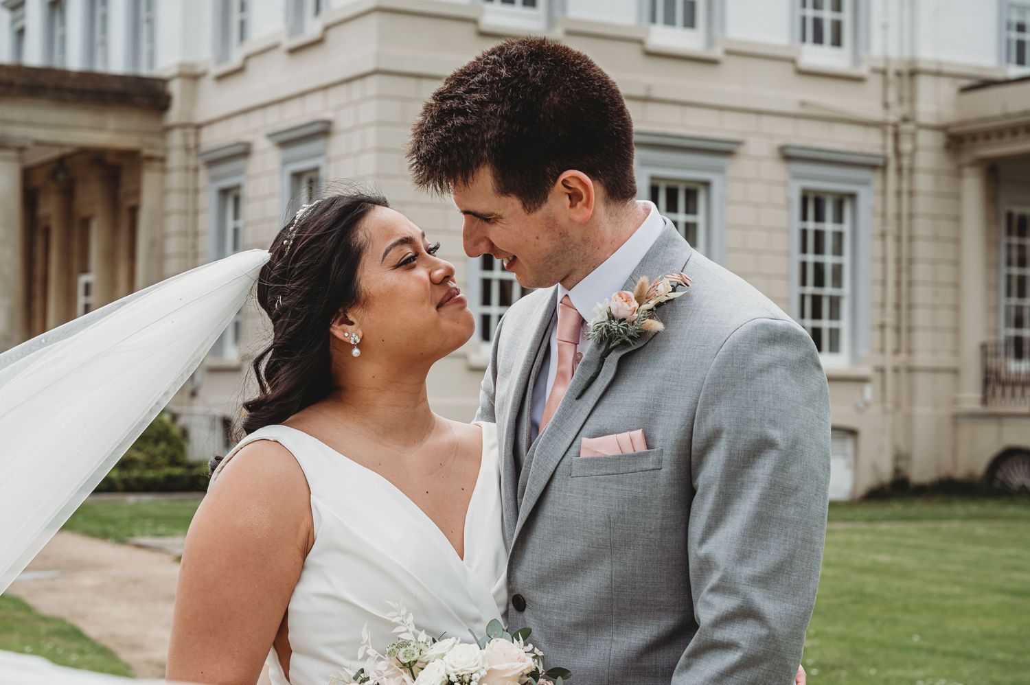 A tender moment between newlyweds in front of a grand estate building, with the bride holding a white floral bouquet.
