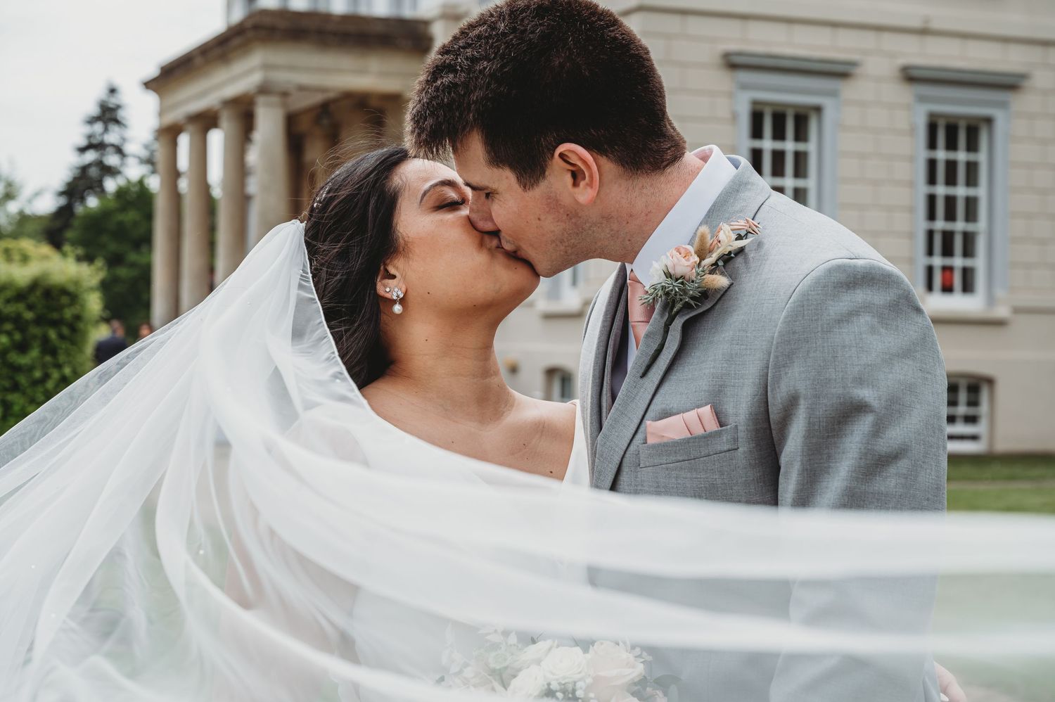 A romantic wedding kiss shared between newlyweds with a flowing white veil and elegant mansion backdrop.