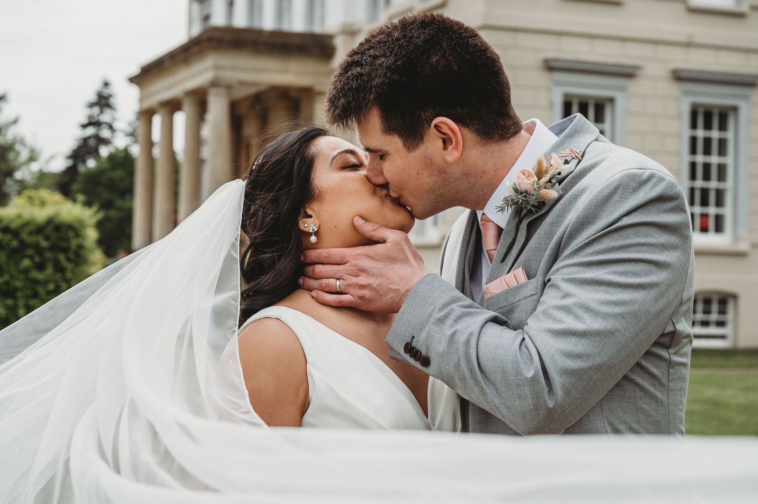 A romantic wedding kiss between newlyweds outside an elegant manor house with flowing white veil in the breeze.