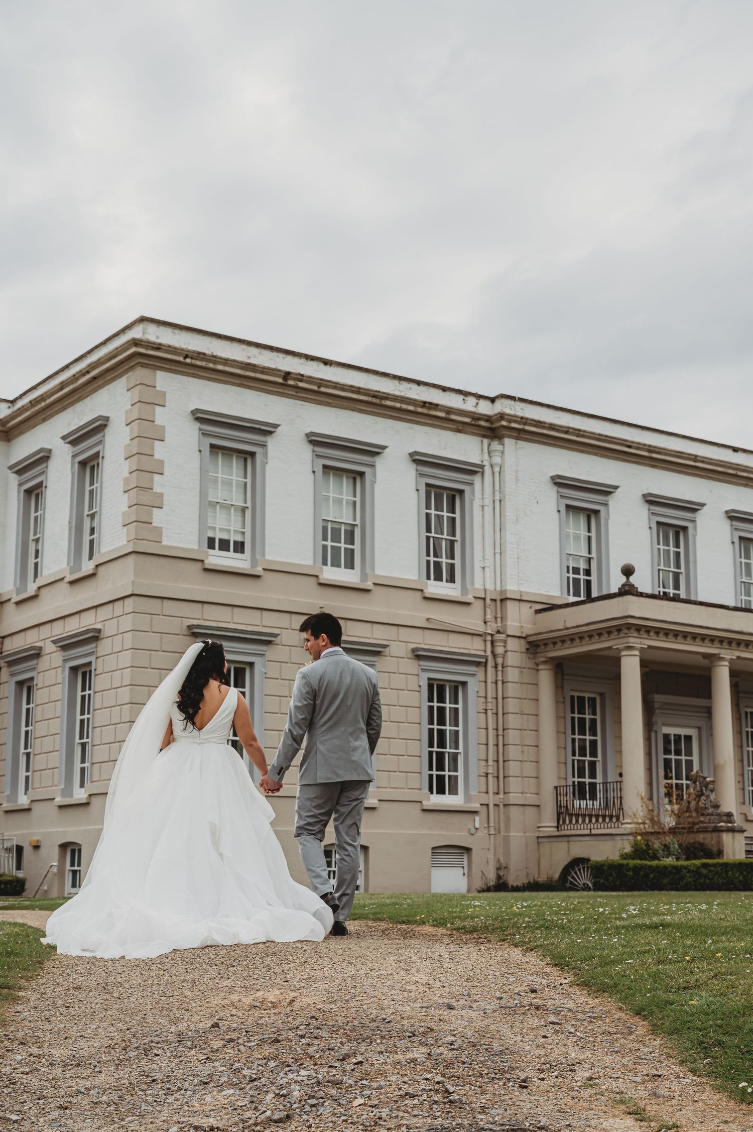 Newlywed couple walking hand in hand on a path leading to a grand historic mansion with white columns and Georgian architecture.