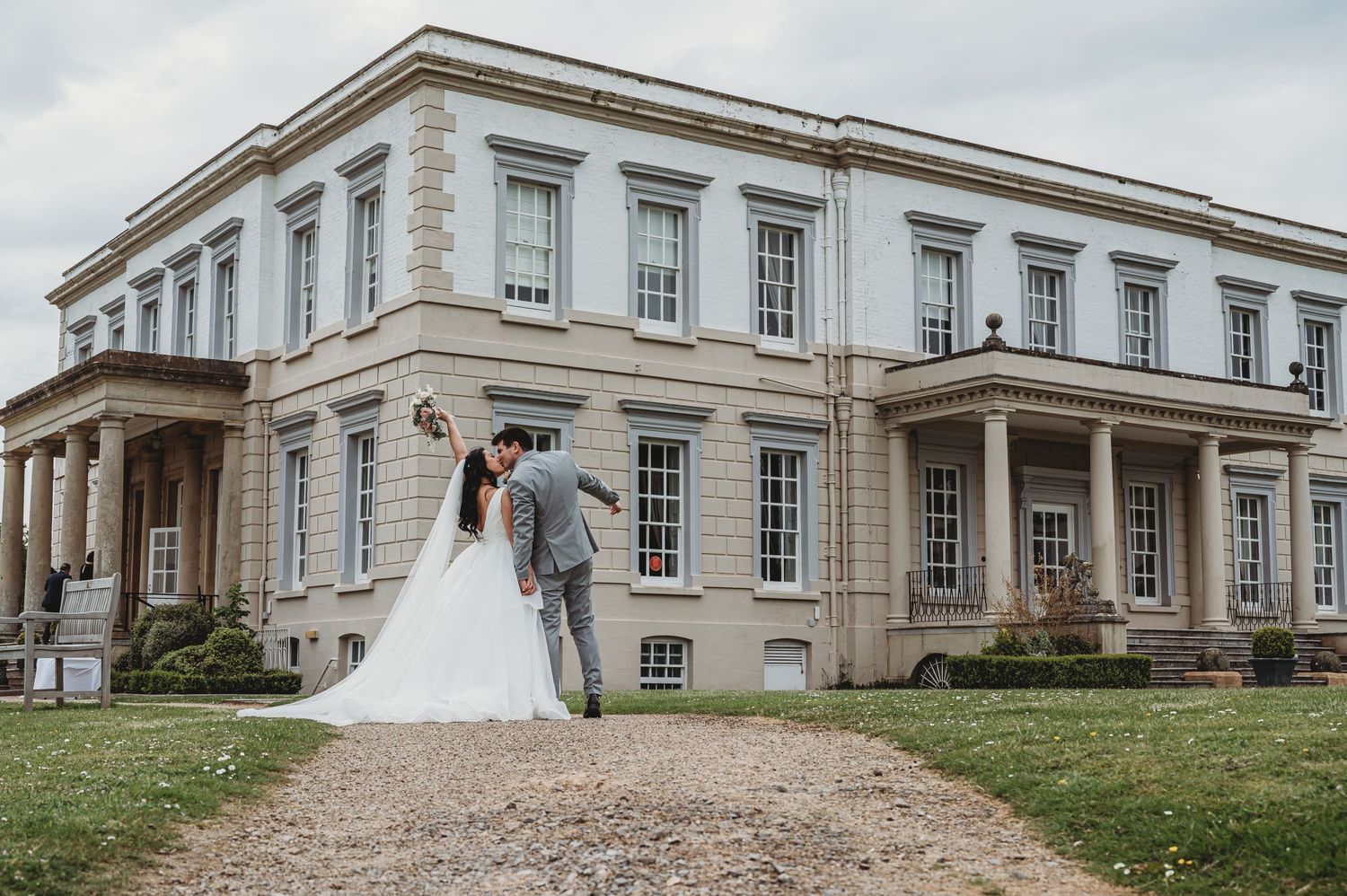 A romantic wedding photo in front of an elegant mansion with classic columns and white facade on a cloudy day.
