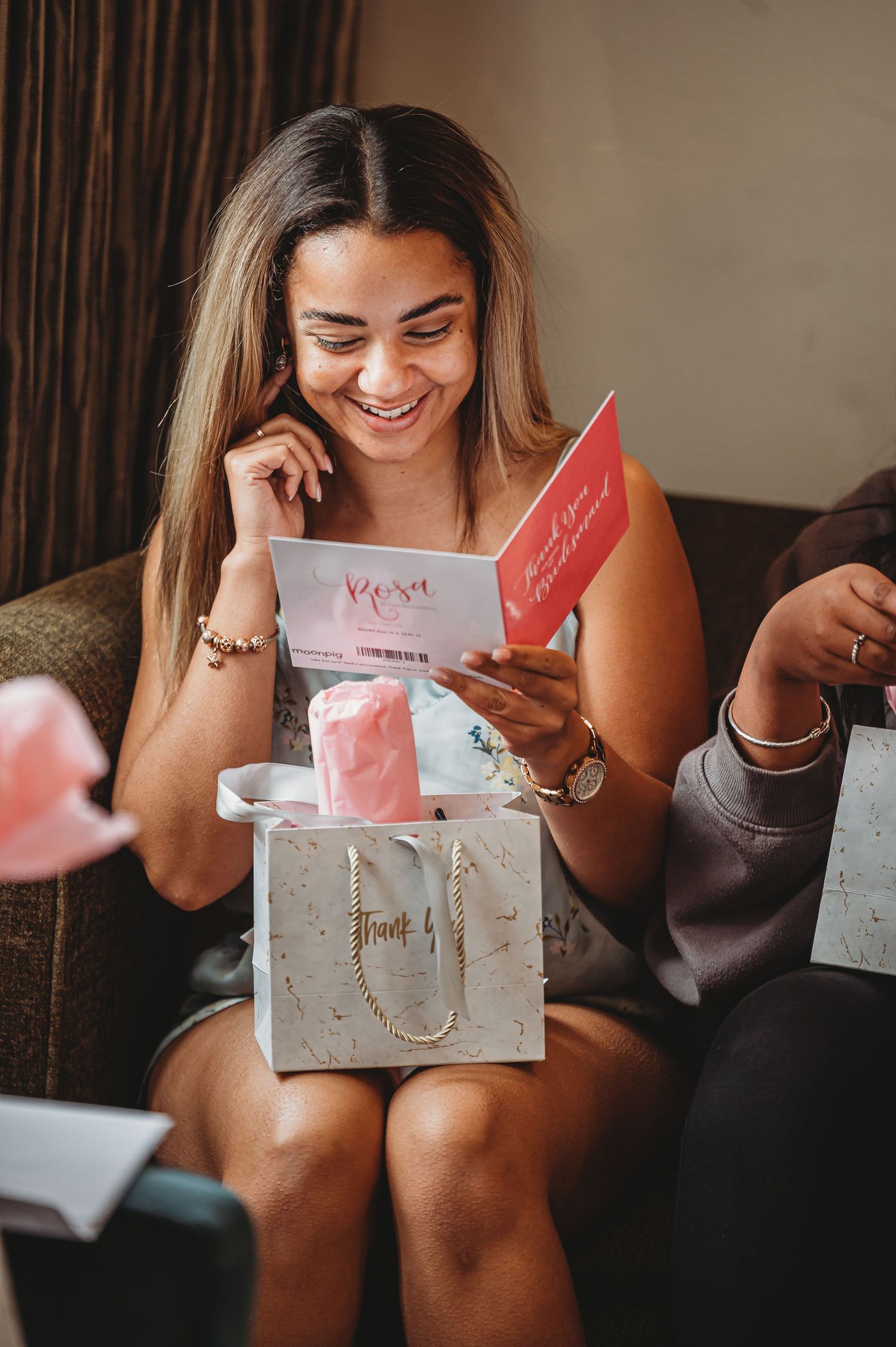 A smiling woman opens a pink gift card while sitting on a couch with a white gift bag nearby.